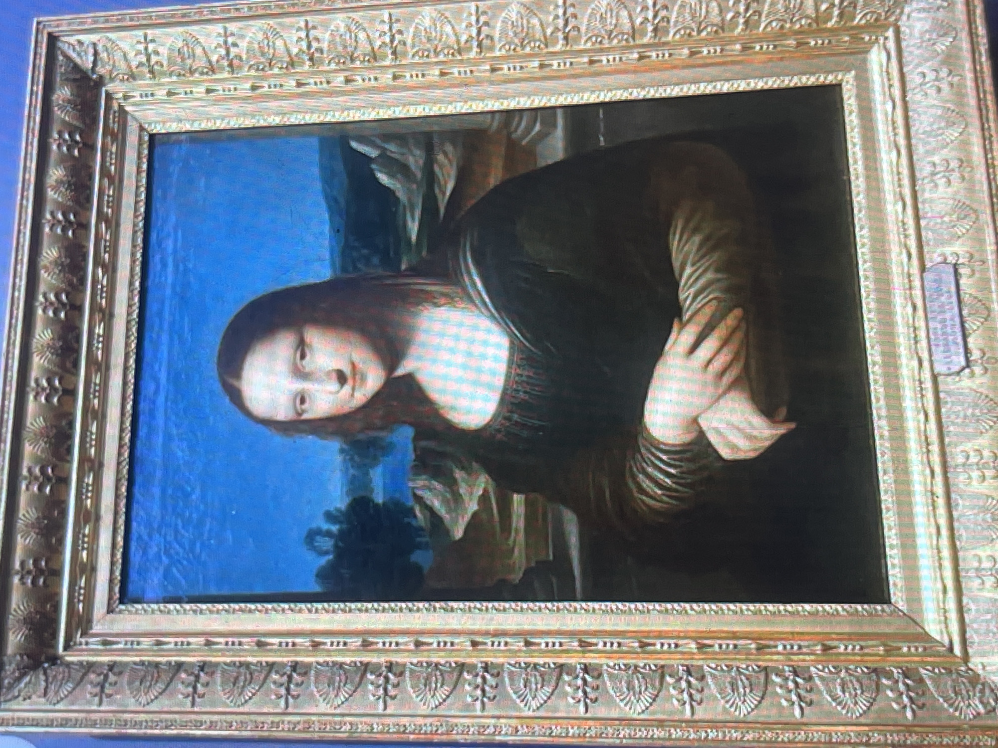 The image shows a framed painting of the Mona Lisa. The painting is an oil on poplar panel by Leonardo da Vinci. The subject is a woman with dark hair and a gentle smile, seated in front of a landscape. The frame is ornate and gold-colored, with intricate carvings. The painting is displayed in a museum or gallery setting, as suggested by the lighting and the presence of what appears to be a plaque below the painting, though the text on the plaque is illegible. The overall impression is of a famous artwork being viewed by an unseen audience. The location in Bishkek City, Kyrgyzstan, is not discernible from the image itself.