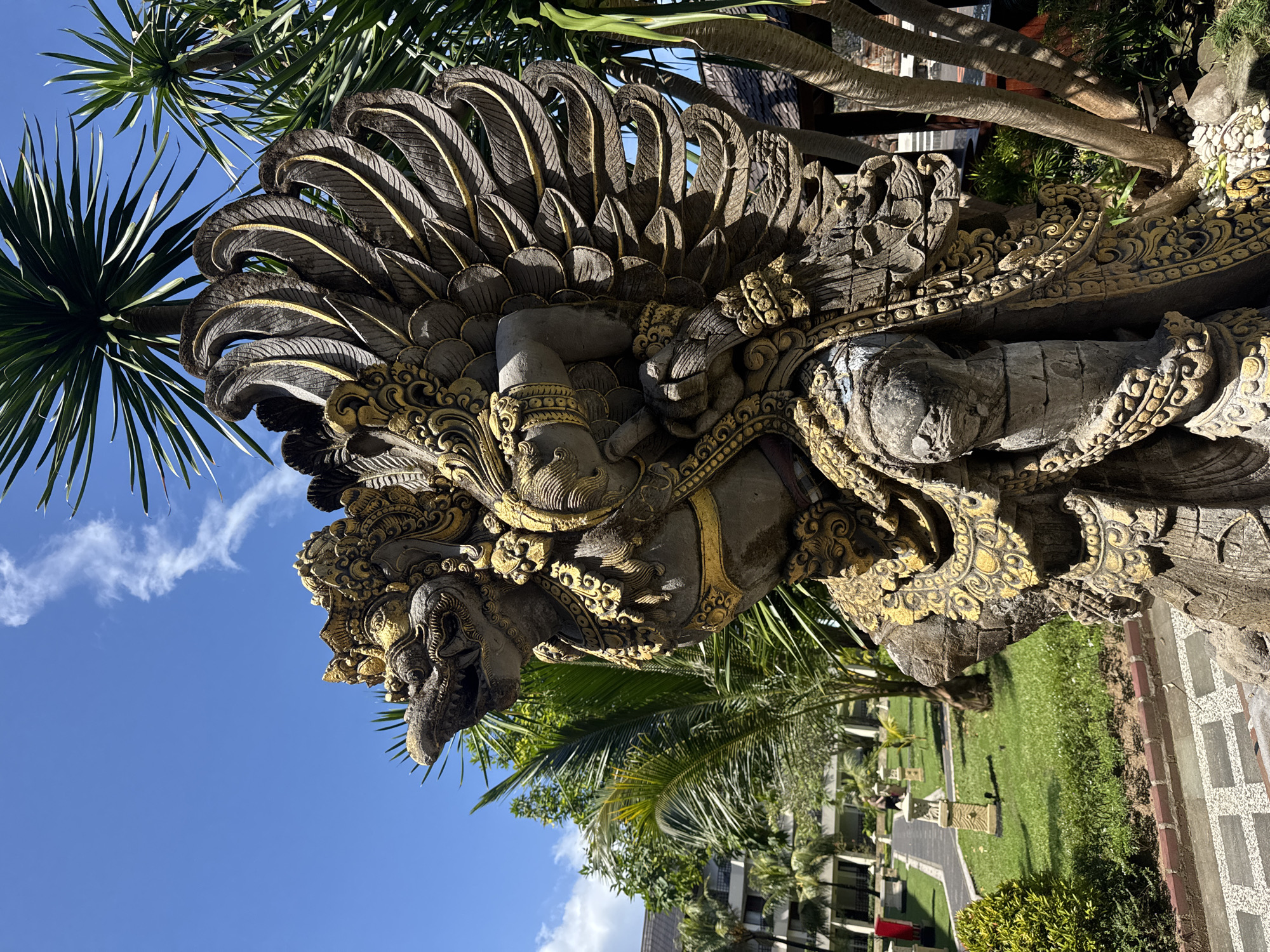 The image depicts a stone statue of a deity, likely Garuda, with intricate carvings and gold-colored accents. The statue is set within a tropical landscape with palm trees and a blue sky with scattered clouds visible in the background. The statue is likely located in a garden or courtyard within the city of Denpasar, Indonesia, as suggested by the Balinese-style architecture and lush vegetation. The statue faces to the right and upward from the perspective of the image.