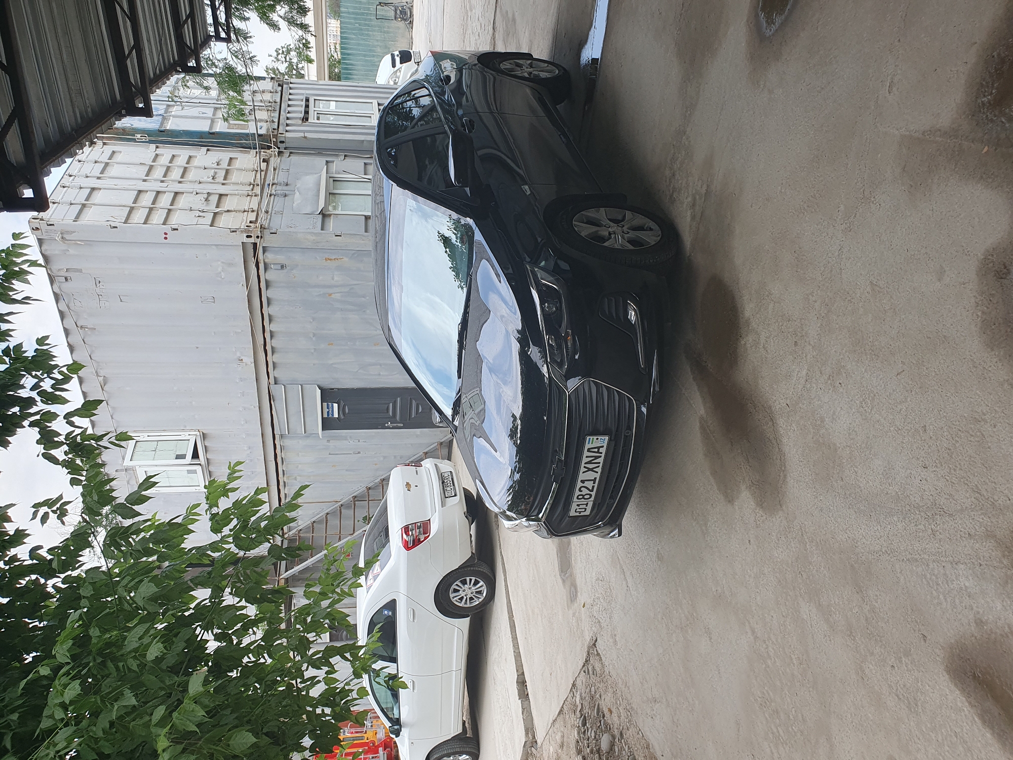 In the city of Tashkent, Uzbekistan, on a concrete-paved area, two Chevrolet sedans are parked. The foreground features a black Chevrolet sedan with license plate "01 821 XNA". Behind it, a white Chevrolet sedan with license plate "GLC 895RC" is visible. In the background, two stacked shipping containers form a temporary structure, with stairs leading to an upper level. The area appears to be an outdoor workspace or storage yard, with hints of a covered area over the black sedan. Green foliage from trees hangs into the frame from the left. The sky is overcast.