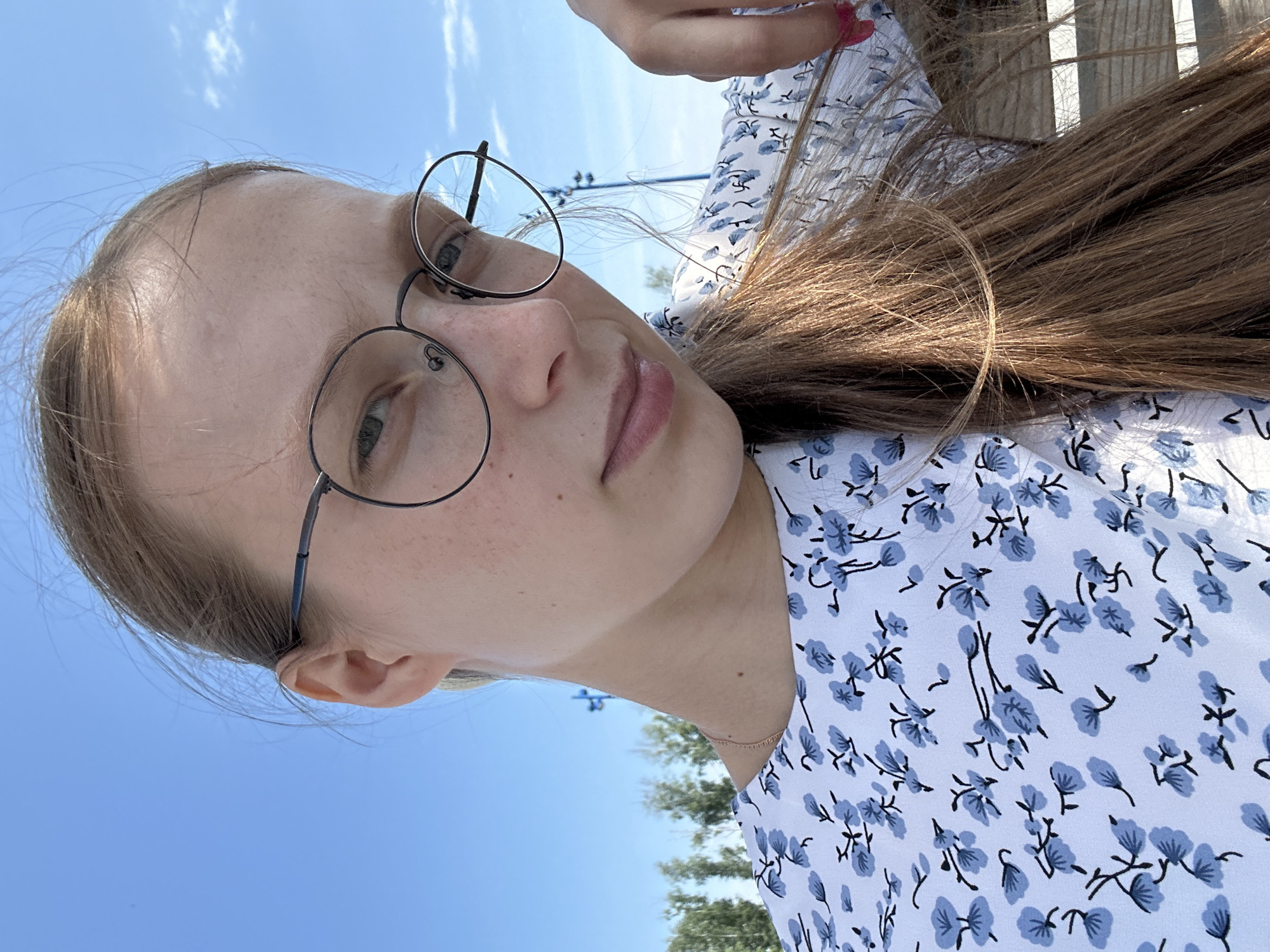 A young woman with long, brown hair is taking a selfie outdoors. She is wearing round, dark-rimmed glasses and a white dress with a blue floral pattern. Her hair is pulled back into a ponytail and falls over her shoulder. She is looking at the camera with a slight smile. The background shows a bright blue sky with a few clouds, and some green trees and structures are visible in the distance. The location appears to be a park or open area, possibly on the outskirts of the city.