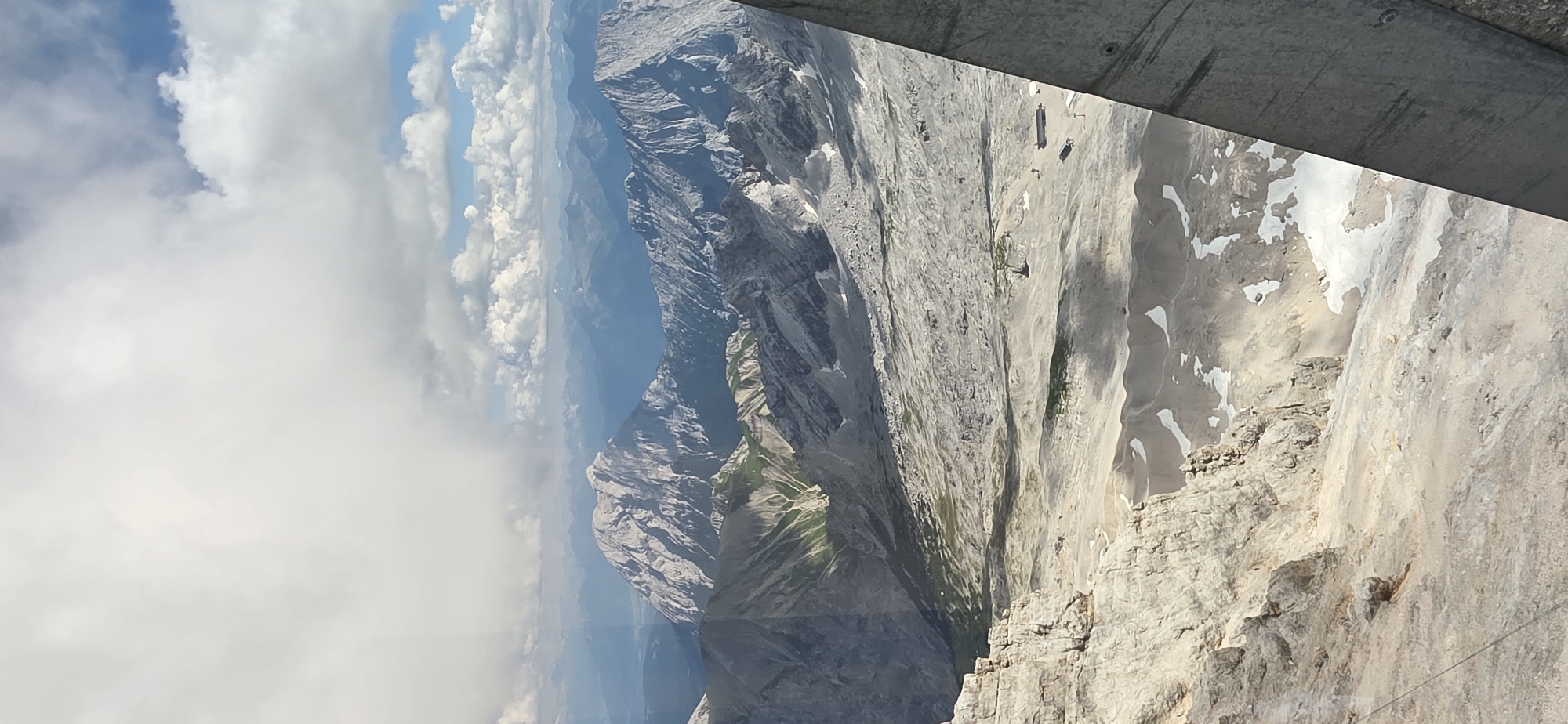 This is a vertical, high-angle photograph of a vast mountain landscape, likely taken from an elevated viewpoint such as a cable car or a mountain peak. In the foreground, on the left, is a jagged, light-colored rocky cliff face. To the right, a dark, angled structural element, possibly part of a building or railing, partially obscures the view.
The midground reveals a wide, undulating expanse of rocky terrain, with patches of snow and sparse green vegetation. Several small structures, appearing to be buildings or facilities, are visible in this area, along with what might be ski lifts or cable car infrastructure. The ground is uneven and rocky, with visible shadows cast by the surrounding mountains.
The background is filled with layers of mountain ranges receding into the distance. The mountains vary in color from light gray to blue-gray, indicating atmospheric perspective. The peaks are rugged and appear to be covered in scree and rock. The sky above is a mix of white, fluffy clouds and patches of bright blue sky, suggesting a partly cloudy day. The overall impression is one of a grand, rugged alpine environment.
The image's perspective and the presence of what appears to be the Zugspitze massif suggest a location in or near Grainau, Germany, specifically at a high elevation. The specific structures and the vastness of the mountain range point towards the area around the Zugspitze, Germany's highest peak. There are no people clearly visible in the image.