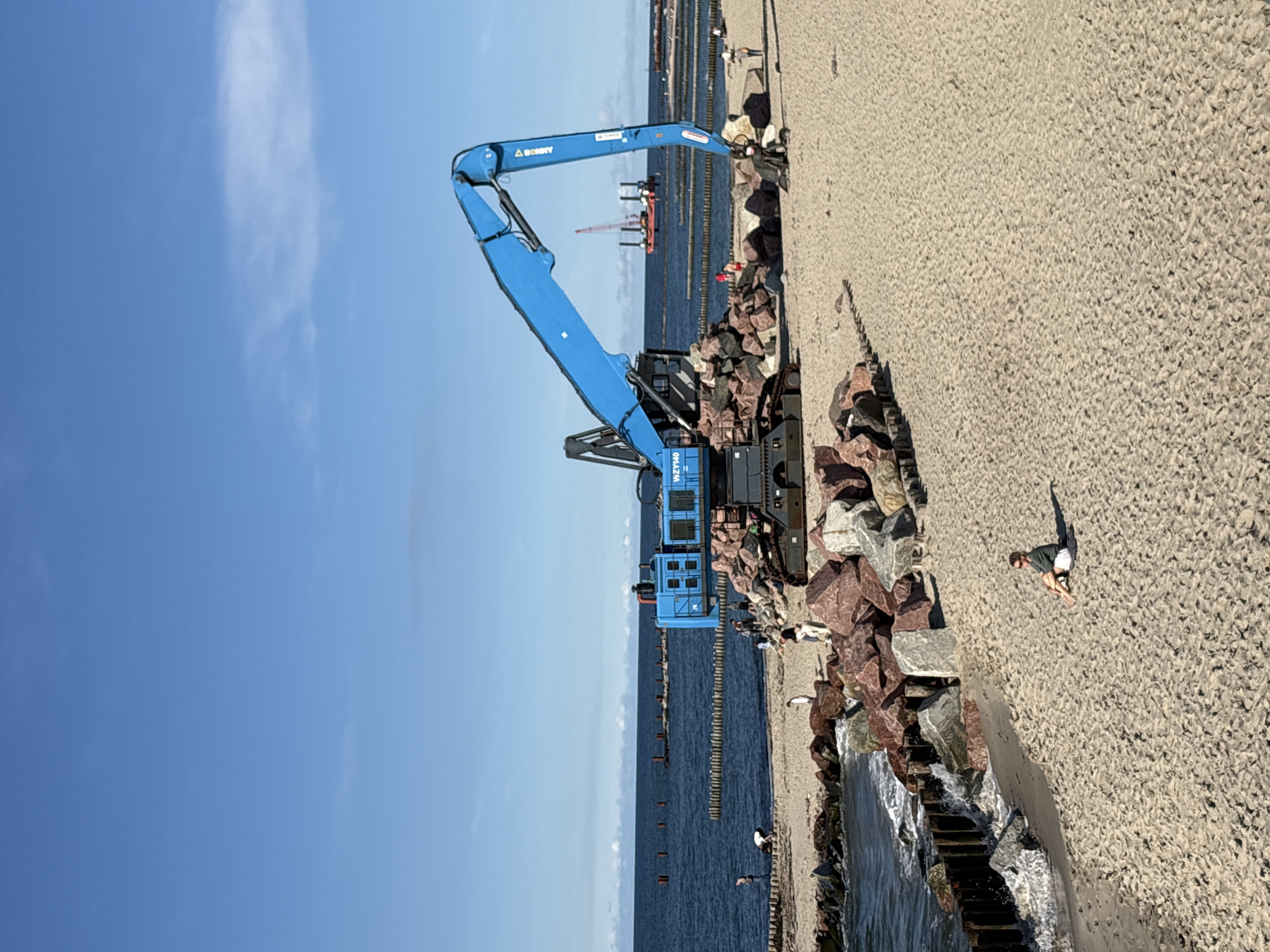 The image depicts a construction scene on a beach, likely in Svetlogorsk, Russia, given the context. A large, bright blue excavator is the central subject, positioned on a sandy beach amidst a pile of large red and gray rocks. The excavator's arm is extended upwards and to the right, over the water. In the background, the calm, blue sea stretches to the horizon, with a clear sky above. Several wooden breakwaters are visible in the water, extending from the shore. Further out at sea, a red and white barge with a crane is anchored. A few people are scattered on the beach. One person is sitting alone on the sand in the foreground, facing away from the camera. Other figures are near the excavator and along the shoreline, some appearing to be observing the construction or engaged in activities on the beach. The overall environment is a sunny day at the seaside, with evidence of ongoing civil engineering work.