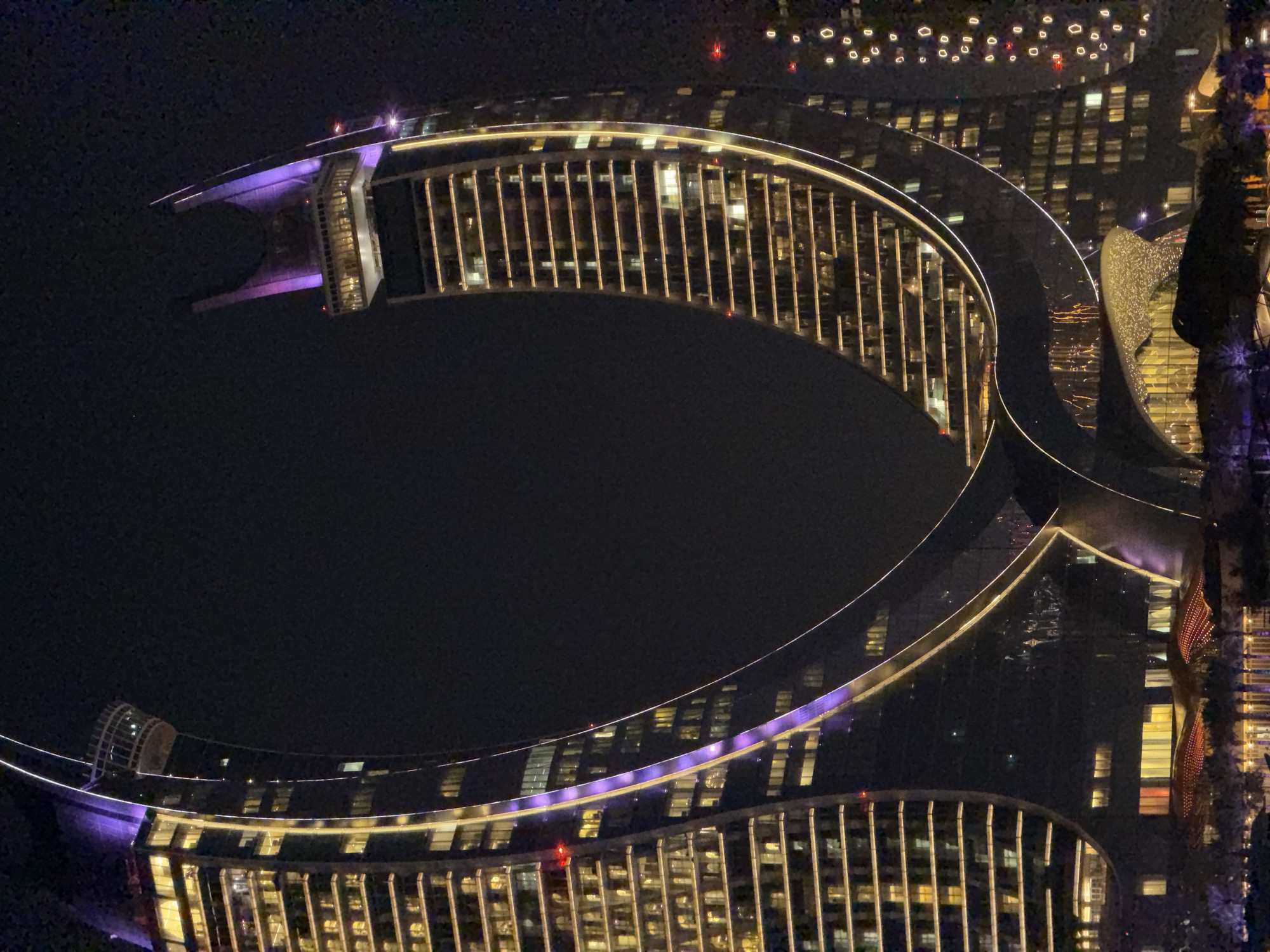 This is an aerial nighttime photograph of a modern, illuminated building complex. The building features a distinctive curved architecture with multiple levels and large windows. The exterior is adorned with dramatic lighting, including purple accents on some sections and warm yellow lights highlighting the architectural details and interior spaces. The overall design suggests a luxurious resort or a high-end commercial establishment.
The scene is set against a dark night sky, with the city lights of a distant urban landscape visible in the background. These distant lights are small and numerous, suggesting a sprawling city. The building itself is the primary subject, its illuminated structure creating a striking contrast with the surrounding darkness.
There are no visible people in the image, but the illuminated windows suggest that the building is occupied. The absence of weather-related details implies clear conditions. The focus is entirely on the architectural magnificence of the building and its interplay with light. The photograph is taken from a high vantage point, giving the viewer an expansive and impressive perspective. The overall mood is one of grandeur and sophistication, highlighting the architectural artistry and the captivating atmosphere of the illuminated building at night.