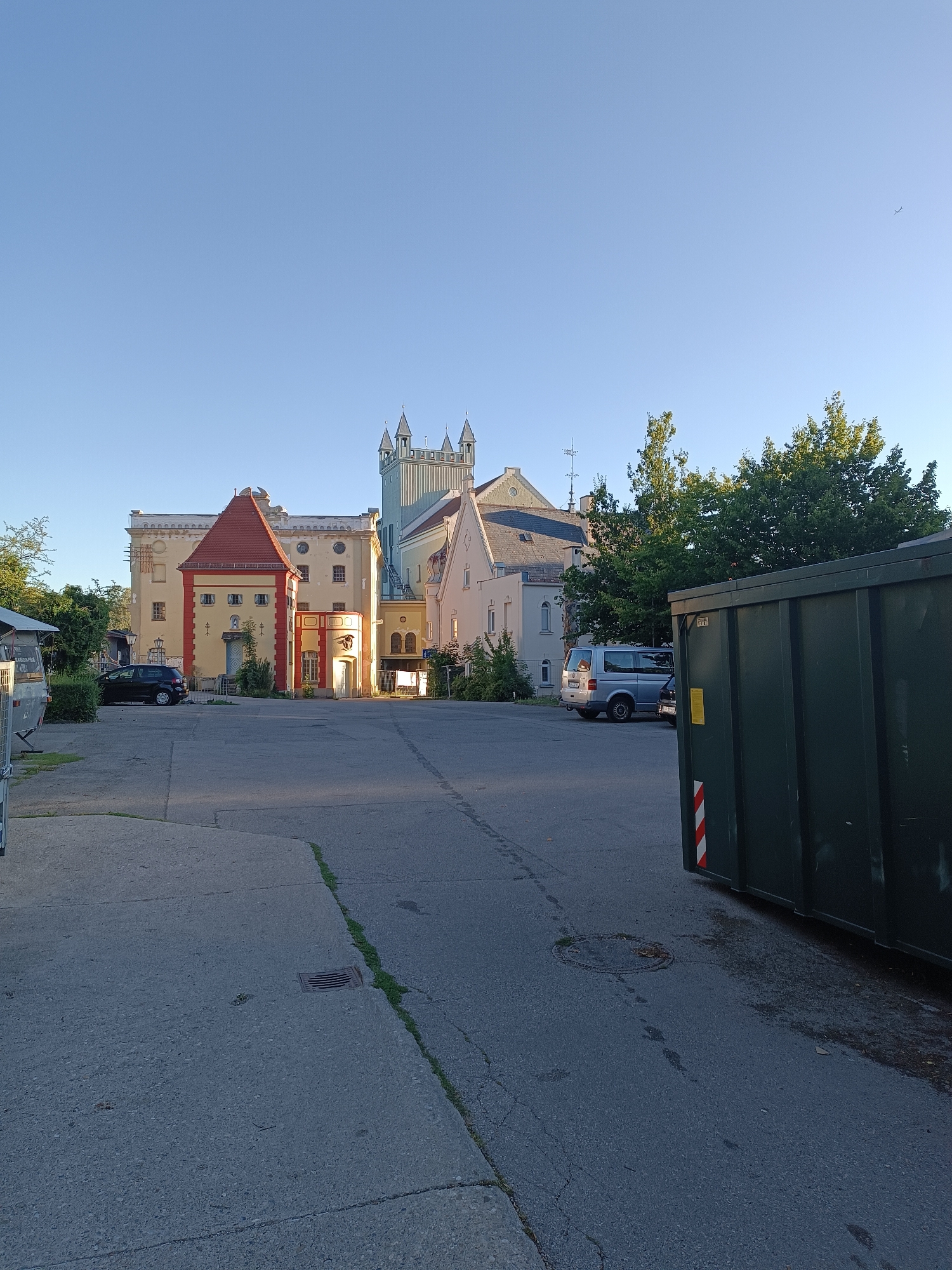 The image depicts an outdoor scene in the city of Fürstenfeldbruck, Germany, on a clear, sunny day. In the foreground, a dark asphalt surface is visible, with a section of lighter concrete to the left, featuring a drain grate. A large, dark green dumpster with red and white reflective stripes is positioned on the right side of the frame. Behind the dumpster, a paved area with some debris and puddles is present.
Dominating the background is a complex of buildings with architectural features reminiscent of a castle or historical structure. The main building is predominantly light yellow, with a prominent red-tiled, steeply pitched roof on a smaller annex on the left. This annex has red trim around its windows and entrance. Further to the right, a taller, pale blue tower with spired turrets rises above the surrounding structures. Other sections of the buildings are painted in lighter hues, with various window shapes and rooflines.
Several vehicles are present: a black hatchback is parked to the left of the red-roofed annex, and a silver minivan is parked further back, near the side of the light blue tower. Trees with dense green foliage are scattered around the buildings, and the clear blue sky occupies the upper portion of the image. The overall impression is one of a historical site or a repurposed industrial building set in a somewhat industrial or service area, indicated by the presence of the dumpster and paved surfaces.