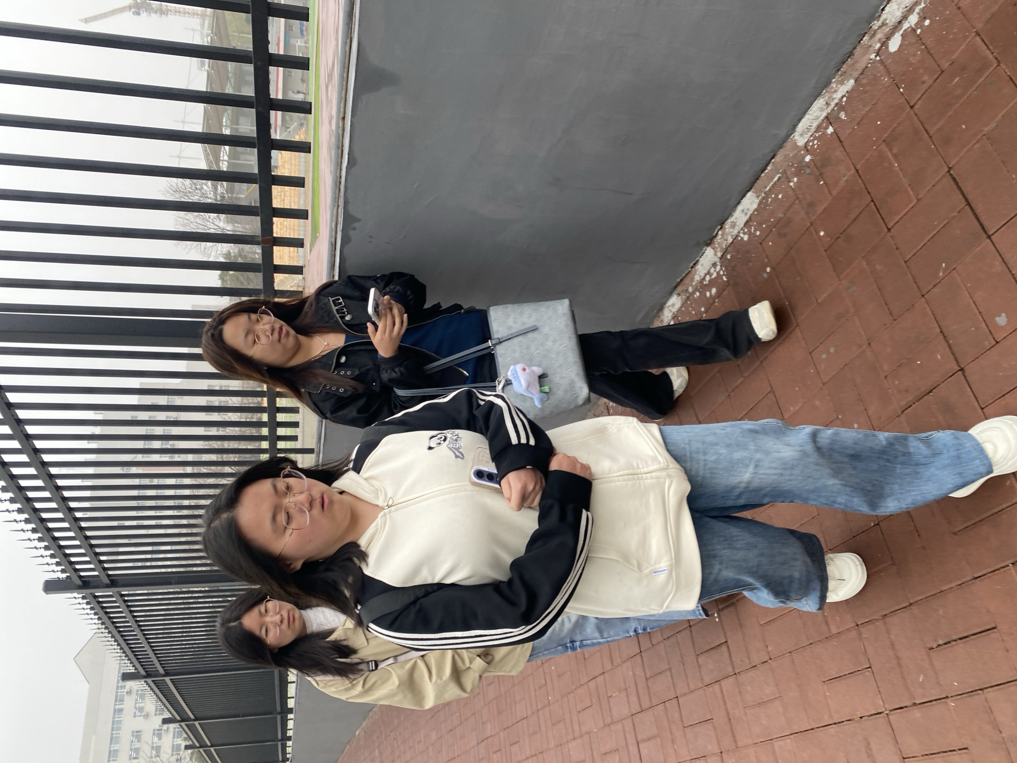 Three East Asian women are sitting on a reddish-brown brick walkway next to a gray wall topped with a black metal railing. The environment appears overcast, typical of a city like Zhongtun, China.
The woman closest to the camera is wearing blue jeans, a white and black hooded jacket, and white sneakers, holding a phone. The second woman behind her is wearing a beige jacket and blue jeans. The third woman is wearing a black leather jacket, a blue shirt, black pants, and white sneakers, and is also holding a phone. A light gray bag sits near her.
The women are positioned parallel to the gray wall and the metal railing. In the background, through the metal railing, buildings and bare trees are visible.