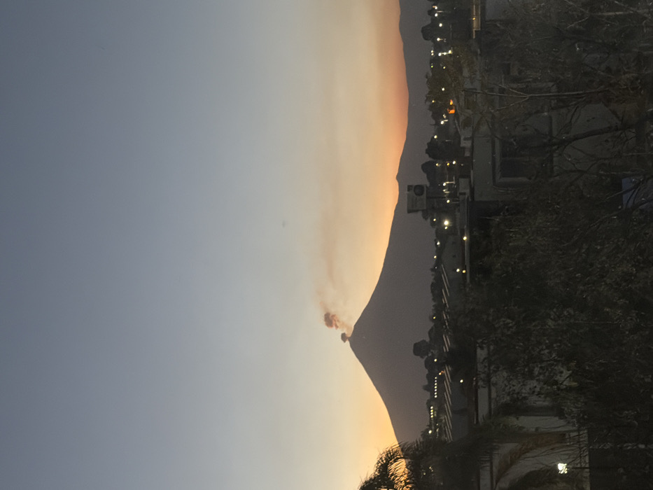 An image shows a mountain at sunset. The sky transitions from blue to orange near the horizon. A plume of smoke rises from the mountain's peak. A road runs along the base of the mountain with streetlights visible. Silhouetted buildings and trees are in the foreground. The image is rotated 90 degrees counter-clockwise.
