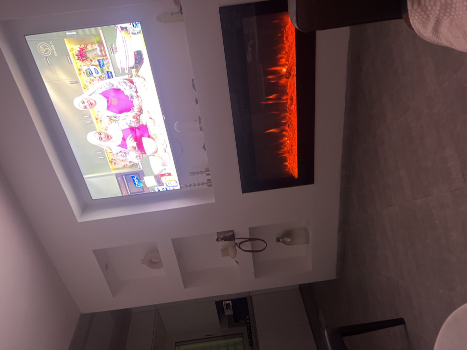 A contemporary living room features a recessed television showing a cooking show with two women wearing hijabs. An electric fireplace with simulated flames is mounted next to the TV. Below the TV and fireplace are built-in shelves displaying decorative items, including candles, vases, and abstract art. The room is modern with tiled flooring and muted lighting. A partial view of furniture is visible in the foreground.
