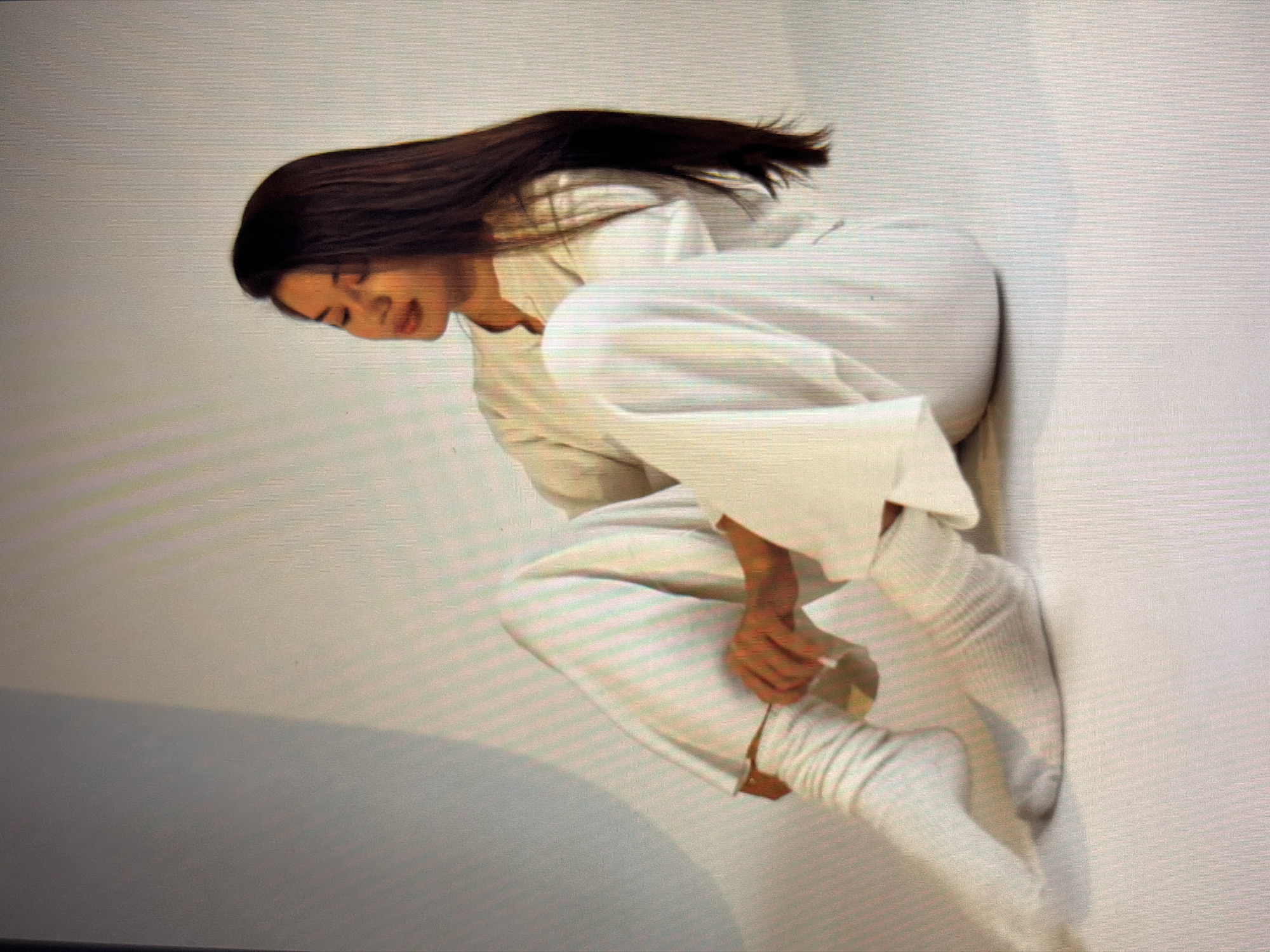 A young woman with long dark hair is sitting with her legs crossed in a studio setting. She is wearing a loose-fitting white outfit consisting of a long-sleeved top and wide-leg pants, with white socks. She is looking down and adjusting her socks with her hands. The background is a plain white studio wall with soft lighting.
