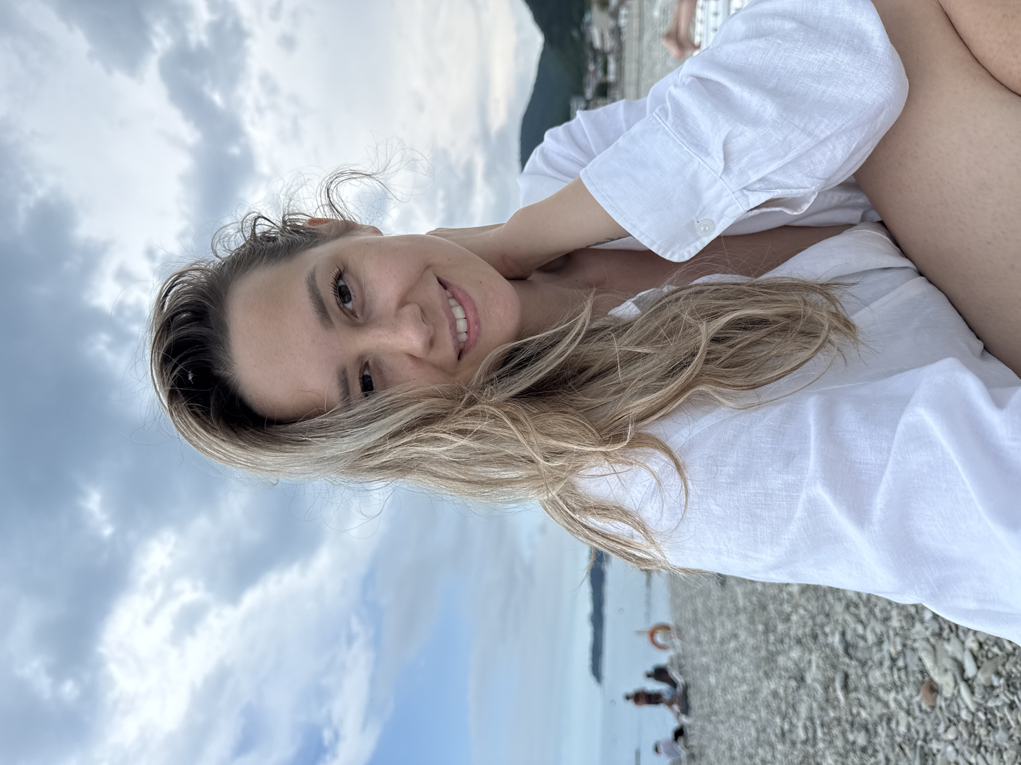 A young woman with blonde hair is sitting on a pebble beach in Nebug, Russia, with the sea and a cloudy sky in the background. She is wearing a white shirt that is unbuttoned, revealing her chest. She is smiling and has her hand on her neck. In the background, there are several people sitting on the beach, and some mountains in the distance.