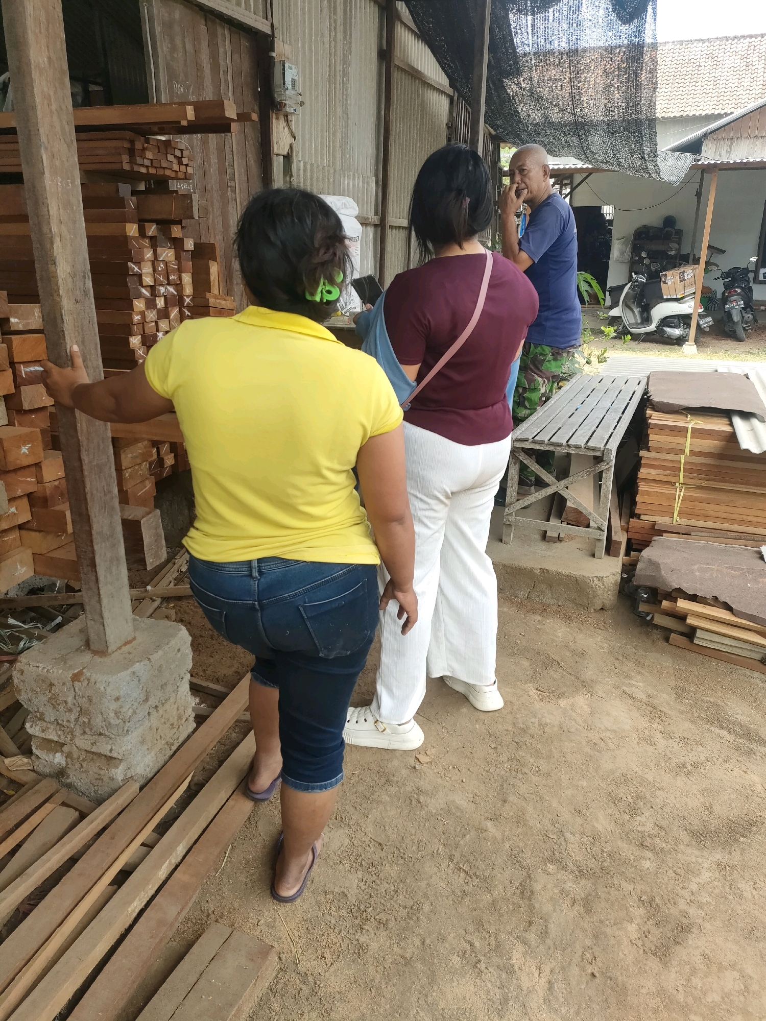 In the city of Balesono, Indonesia, an outdoor scene unfolds at what appears to be a lumber yard or construction supply area. Three individuals are present, with their backs to the camera, suggesting they are engaged in a transaction or discussion.
The focal point is a person wearing a bright yellow t-shirt and dark blue jeans. They are leaning on a large wooden post, their right hand resting on it. Their posture indicates they might be examining the wood or waiting. Behind this person, another individual is visible, dressed in a maroon t-shirt and white pants. A light-colored strap, possibly from a bag, is draped across their chest. The third person, standing further back, is a man in a blue t-shirt and camouflage pants. He has his hand raised to his face, as if speaking or thinking.
The environment is characterized by stacks of lumber, both rough-hewn and processed, arranged in orderly piles. Some of the wood is secured with yellow straps. A weathered wooden bench sits to the right of the individuals, and in the background, a building with a corrugated metal roof and walls is visible, along with two motorcycles and what appears to be a white scooter. The ground is a mixture of dirt and wood shavings, consistent with an outdoor workspace. The overall impression is one of a functional, working environment related to wood products.