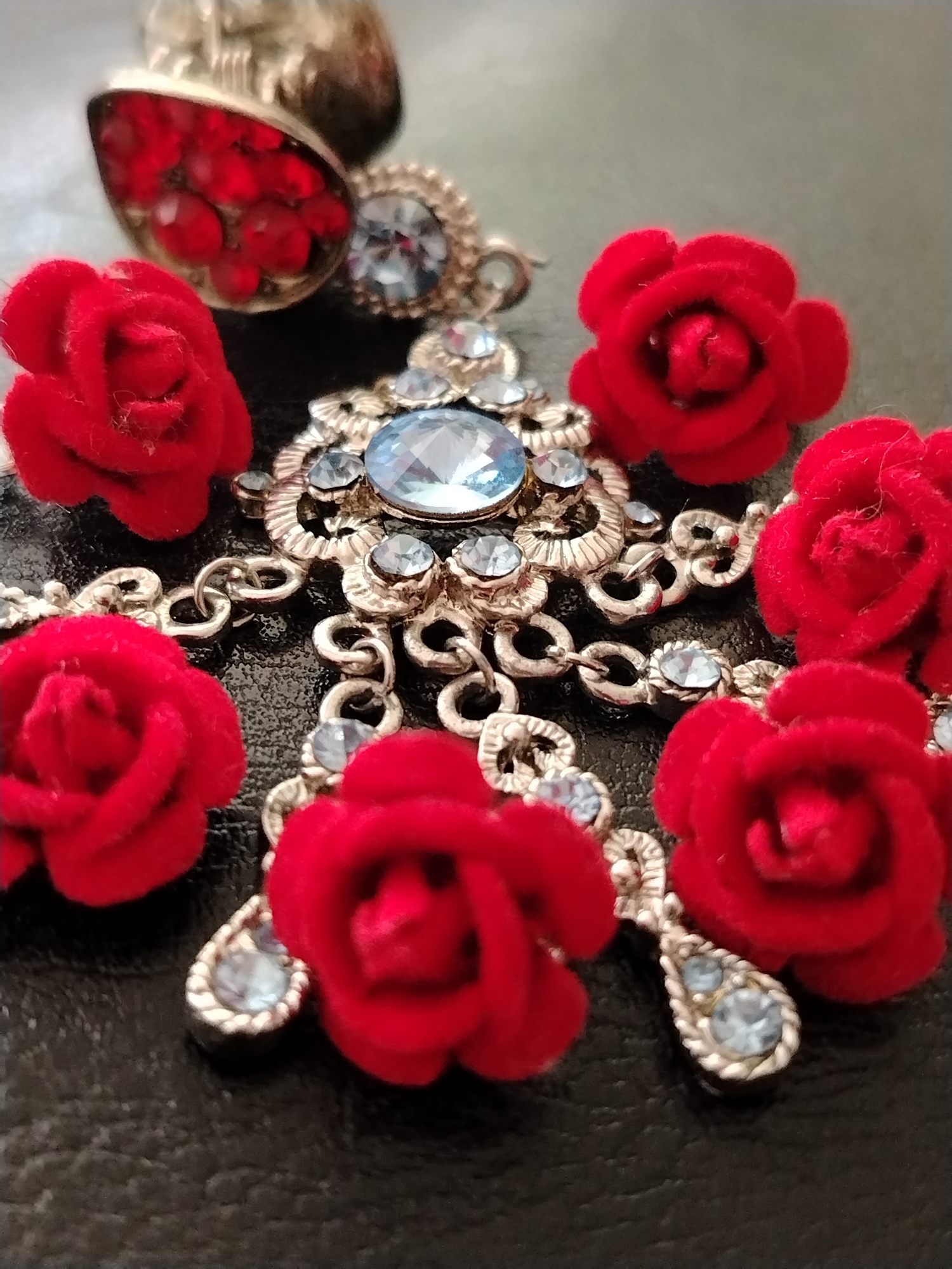 The image displays a close-up of a piece of jewelry on a dark, textured surface. The jewelry features several red velvet roses interspersed with silver-toned metalwork adorned with small, light blue rhinestones. A larger, faceted light blue gemstone is centrally located on a cross-shaped pendant. In the upper left, a distinct teardrop-shaped pendant filled with red stones is visible. The overall aesthetic is ornate and decorative. There is no information in the image to suggest a location in Krasnodar, Russia.