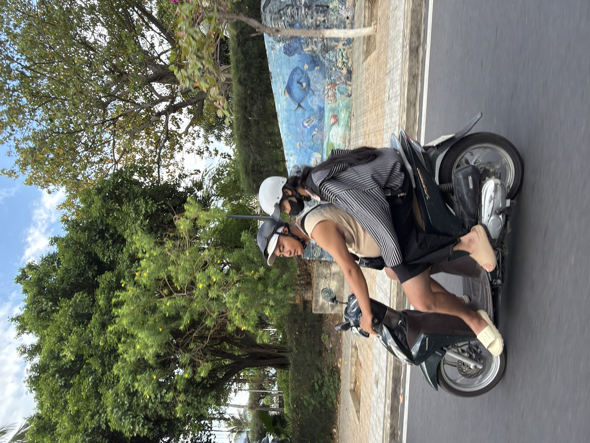 In Nha Trang, Vietnam, two individuals are riding a dark green scooter on a paved road. The male driver in the front wears a tan sleeveless shirt, black shorts, a dark helmet with a white brim, and white slides. The female passenger behind him wears a striped long-sleeved shirt, black pants, a white helmet, and a black face mask. The scooter is in motion, evidenced by the slight blur in the background. To the right of the road is a concrete wall with a mural depicting an underwater scene with fish and coral. Beyond the wall, lush green trees and blue sky with scattered clouds form the background. The sidewalk is made of light-colored square tiles.