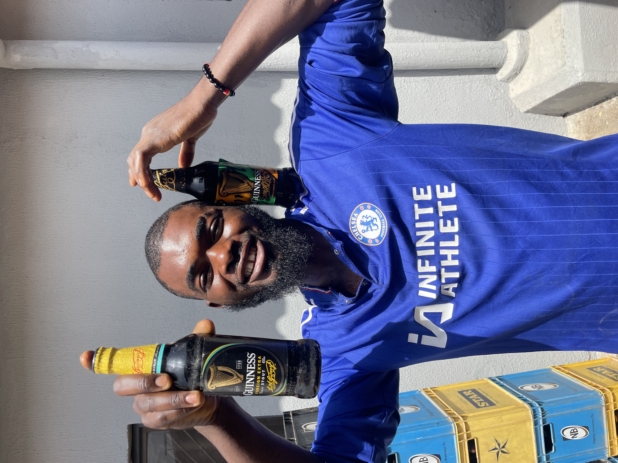 In this media file, a Black man wearing a blue Chelsea Football Club jersey with "INFINITE ATHLETE" printed on it is posing for the camera. He holds two bottles of Guinness Foreign Extra Stout, one in each hand. The man has a beard and is smiling broadly. The scene appears to be outdoors, with a white wall and the sun casting shadows, suggesting it is daytime. In the background, stacks of blue and yellow beer crates, some labeled "STAR" and "NB Nigerian Breweries Plc.", are visible. The overall mood is cheerful and celebratory, possibly indicating a moment of relaxation or enjoyment. The location context is Jos, Nigeria.