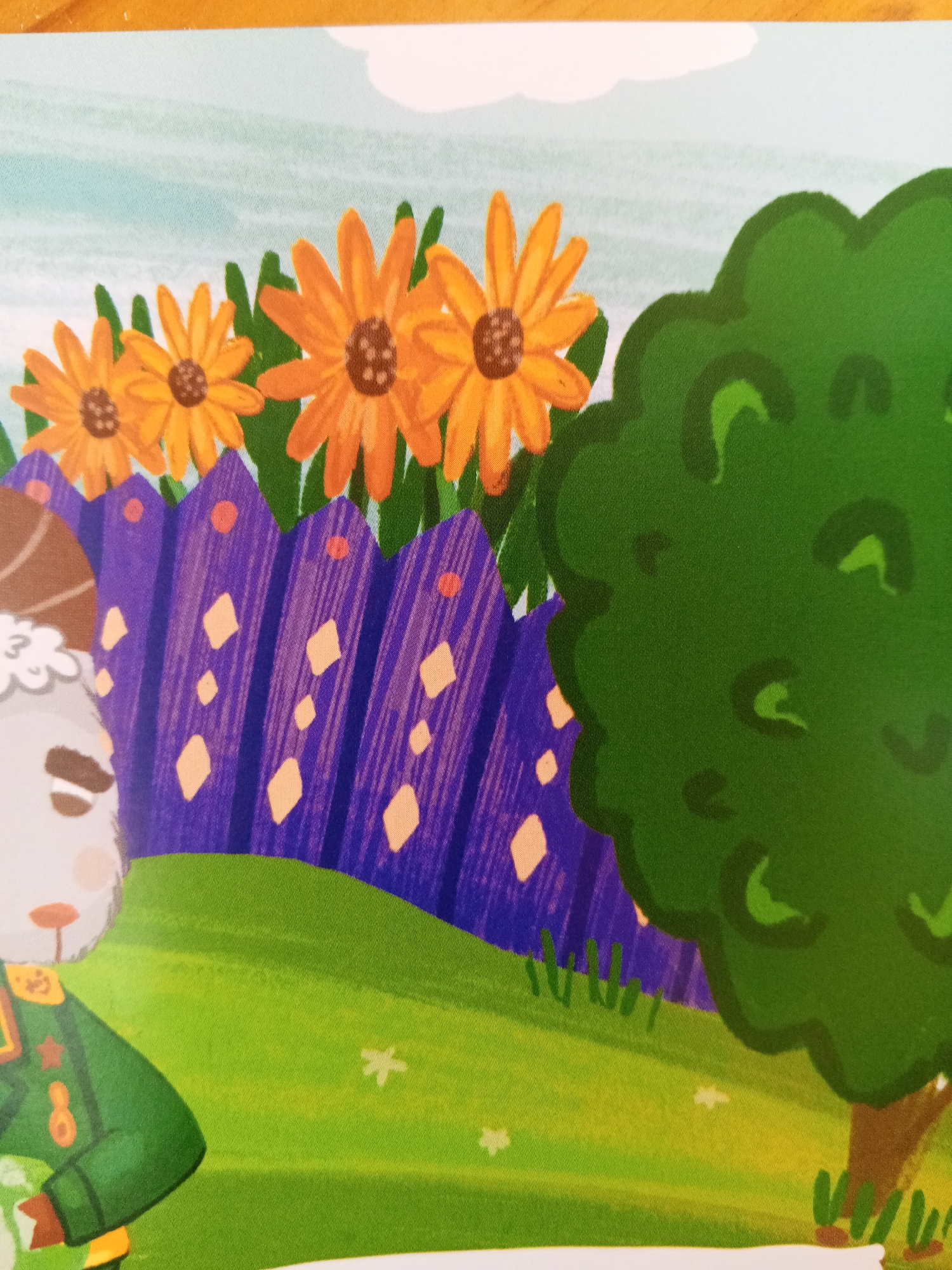 This image depicts a cartoon scene with a light blue sky and a vibrant green landscape. In the foreground, a character resembling a white rabbit or sheep, wearing a green military-style uniform with gold accents and a red star insignia, is partially visible. The character has a neutral expression. Behind the character, a purple picket fence with orange diamond and dot details runs horizontally across the image. Several large, orange daisy-like flowers with brown centers are positioned above the fence. To the right of the scene, a large, dark green, cartoonish tree with a brown trunk stands prominently. The ground is a bright green grassy hill with some small white star-shaped elements scattered on it. The image appears to be a still from an animated production or a children's book illustration. There is no discernible information within the image that can be used to determine its location in Chistopol, Russia.