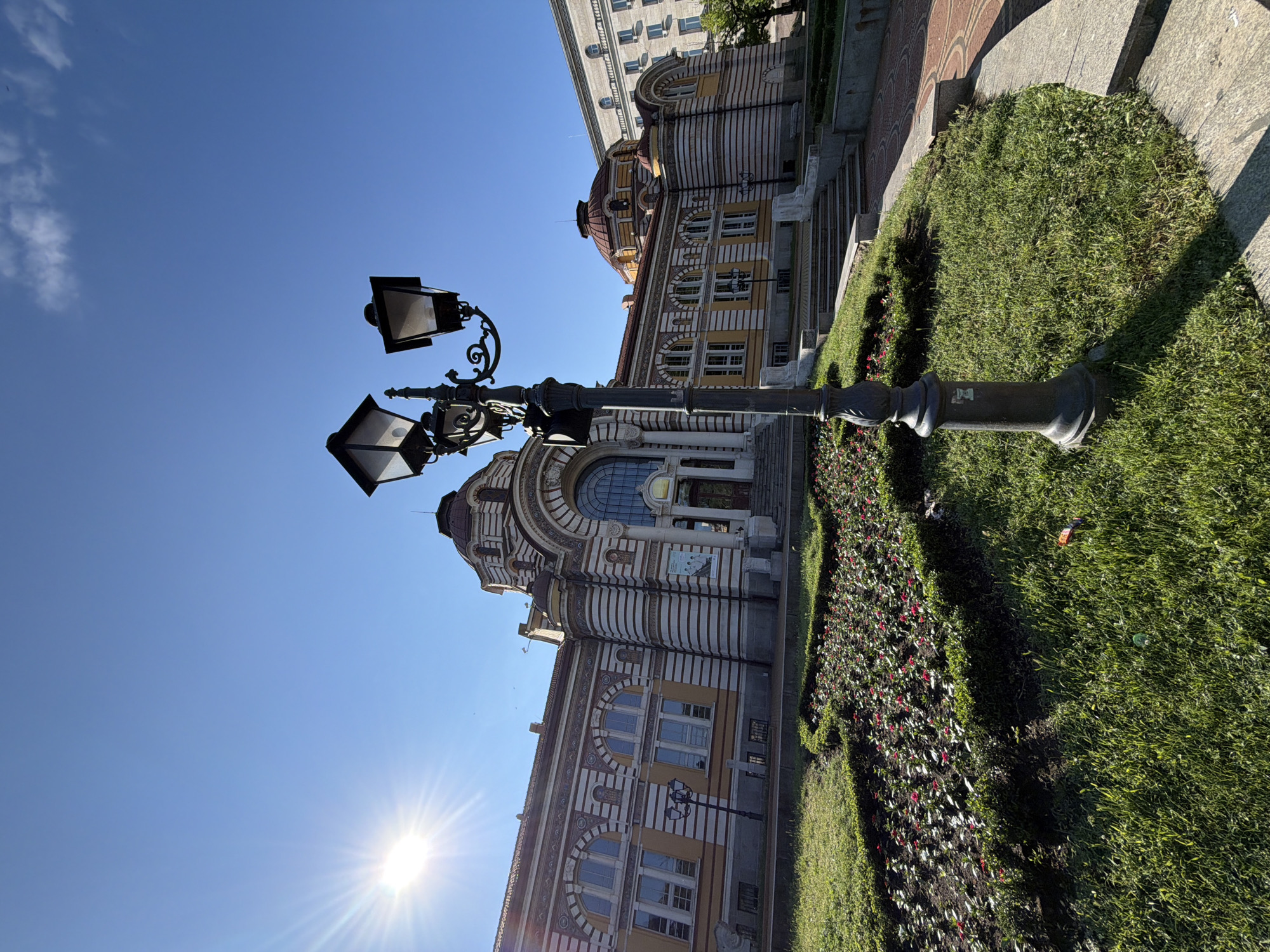 The original image is a wide-angle shot of the Central Mineral Baths in Sofia, Bulgaria, on a clear, sunny day. The building, a prominent architectural landmark, is characterized by its striking yellow and white striped facade, red-tiled domes, and arched windows. In the foreground, a black ornate lamppost stands tall amidst a well-maintained green lawn dotted with flowerbeds featuring red flowers. The sun is bright, casting lens flare effects and creating a stark contrast with the clear blue sky. To the right of the Central Baths, another building with a lighter facade and visible windows can be seen. There are no people visible in the frame. The scene captures a public space, likely an urban park or square, in front of the historic bathhouse. The photograph's perspective emphasizes the grandeur of the architecture and the surrounding greenery under the bright sunlight.