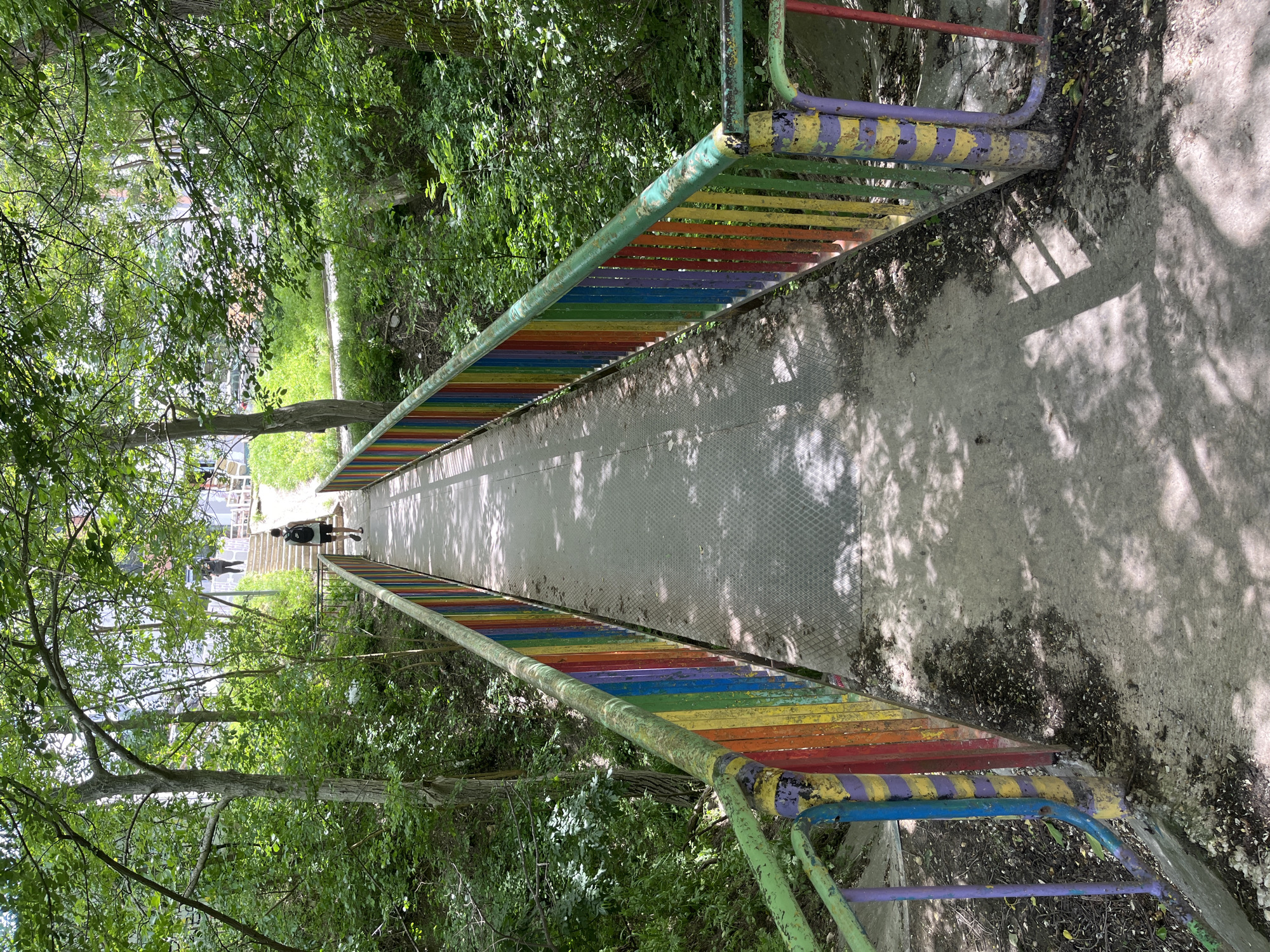 A slightly high-angle, full shot presents a rainbow-colored metal bridge in a park-like setting. The bridge's railings are painted with alternating bands of green, blue, yellow, orange, and red. The bridge surface appears to be concrete with a textured pattern. The background consists of lush green trees and foliage, casting dappled shadows on the bridge. Two human figures are present in the center of the bridge at a distance. It is a park in Varna, Bulgaria.