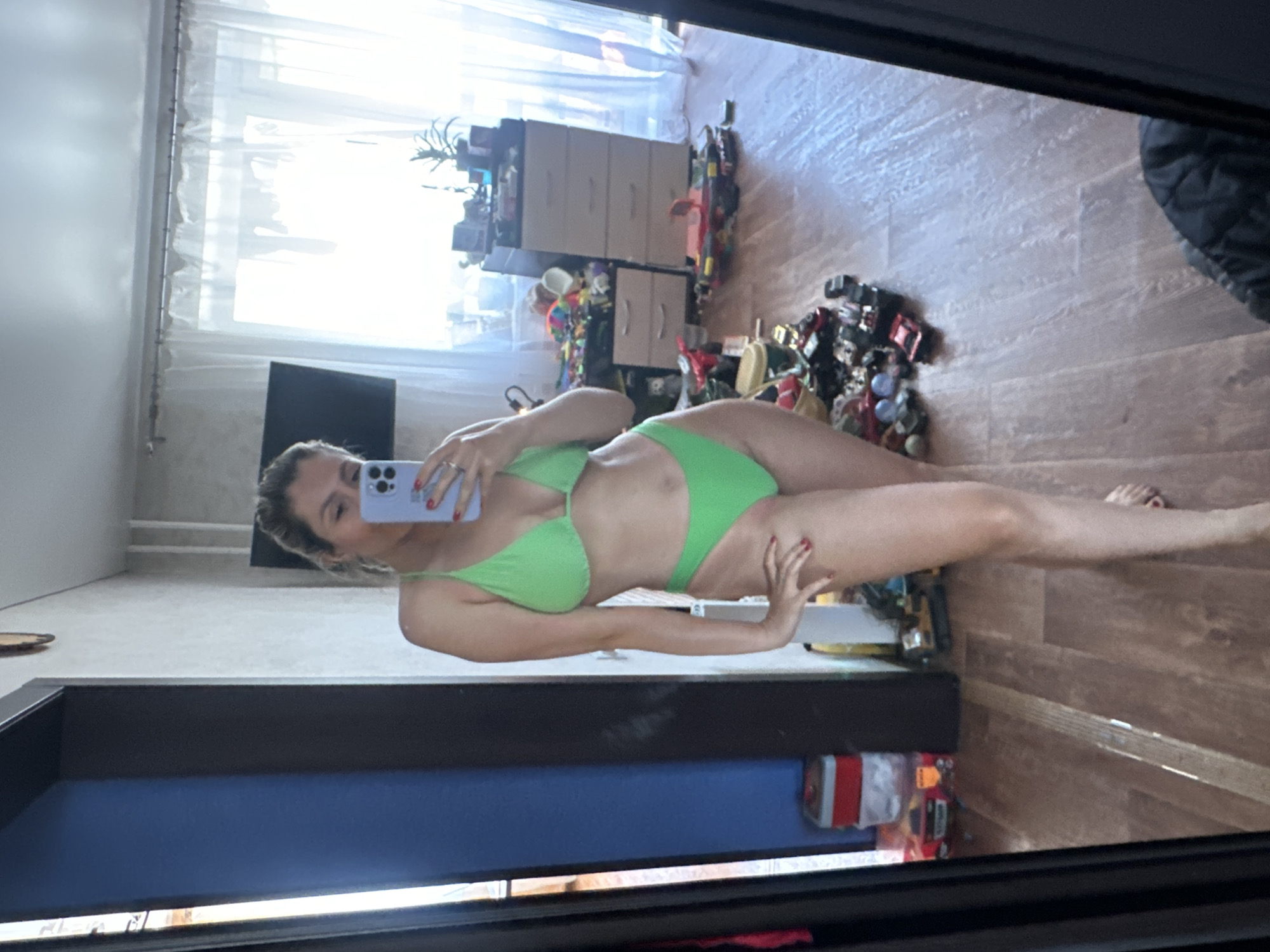 A woman stands in front of a mirror, taking a selfie. She is wearing a bright green bikini. The room appears to be a bedroom, with a dresser, window with sheer curtains, and a closet visible. A collection of toys is scattered on the floor. The image appears to be taken in Novoaltaysk, Russia.