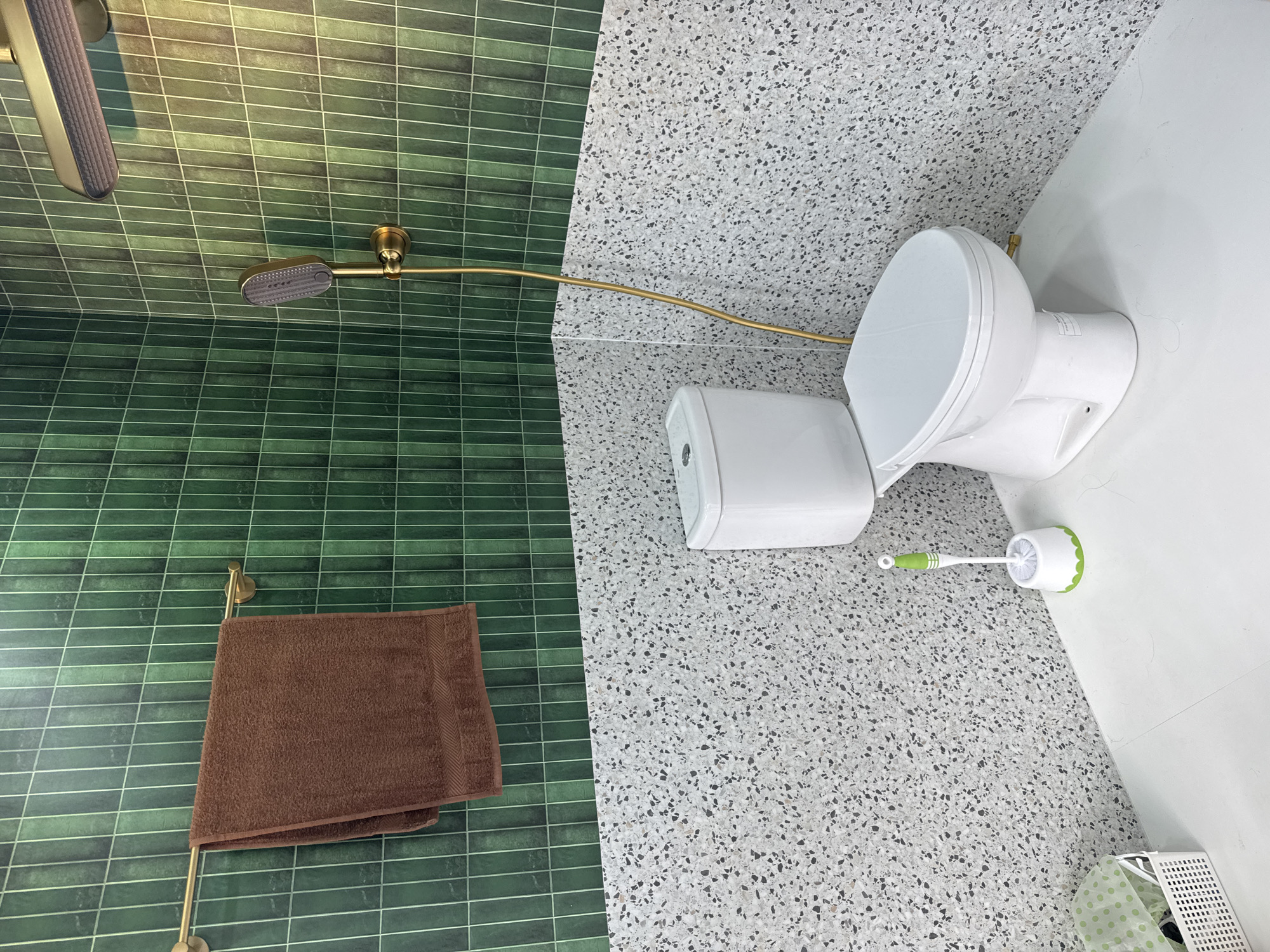 This is an interior shot of a bathroom. The bathroom has two distinct sections based on wall tiling. On the left, the wall is covered in horizontal, green, rectangular tiles with light grout. A brown towel is hanging on a gold-colored towel rack. Above the towel rack, there is a gold-colored shower head attached to a longer, gold-colored pipe extending towards the right. On the right side, the wall has a terrazzo-like pattern with small black and gray specks. A white toilet is positioned here, with its seat closed. To the right of the toilet, there is a toilet brush with a green and white handle and a white bristled brush with green accents. At the bottom right corner, a partial view of a green and white patterned basket is visible. The location is Denpasar, Indonesia.