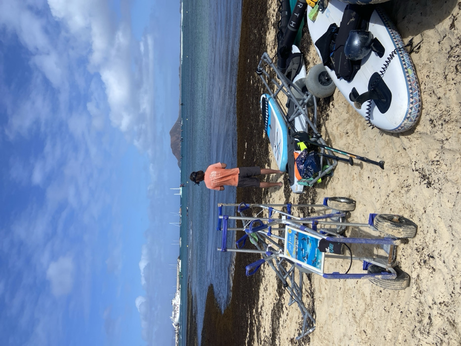 The image shows a sandy beach in Corralejo, Spain (latitude: 28.734, longitude: -13.867). One person is standing on the beach facing away from the camera, while a second person is partially obscured by the cart. The person standing is wearing a pink shirt and blue shorts. The person on the cart is wearing a black shirt and black shorts. There are three carts in the image, and the cart furthest from the camera has a blue and silver frame with an advertisement on it. The other two carts are similar in color and design. The beach is covered in seaweed and a few small boats can be seen in the distance, as well as a large mountain in the background. The sky is blue with white clouds.