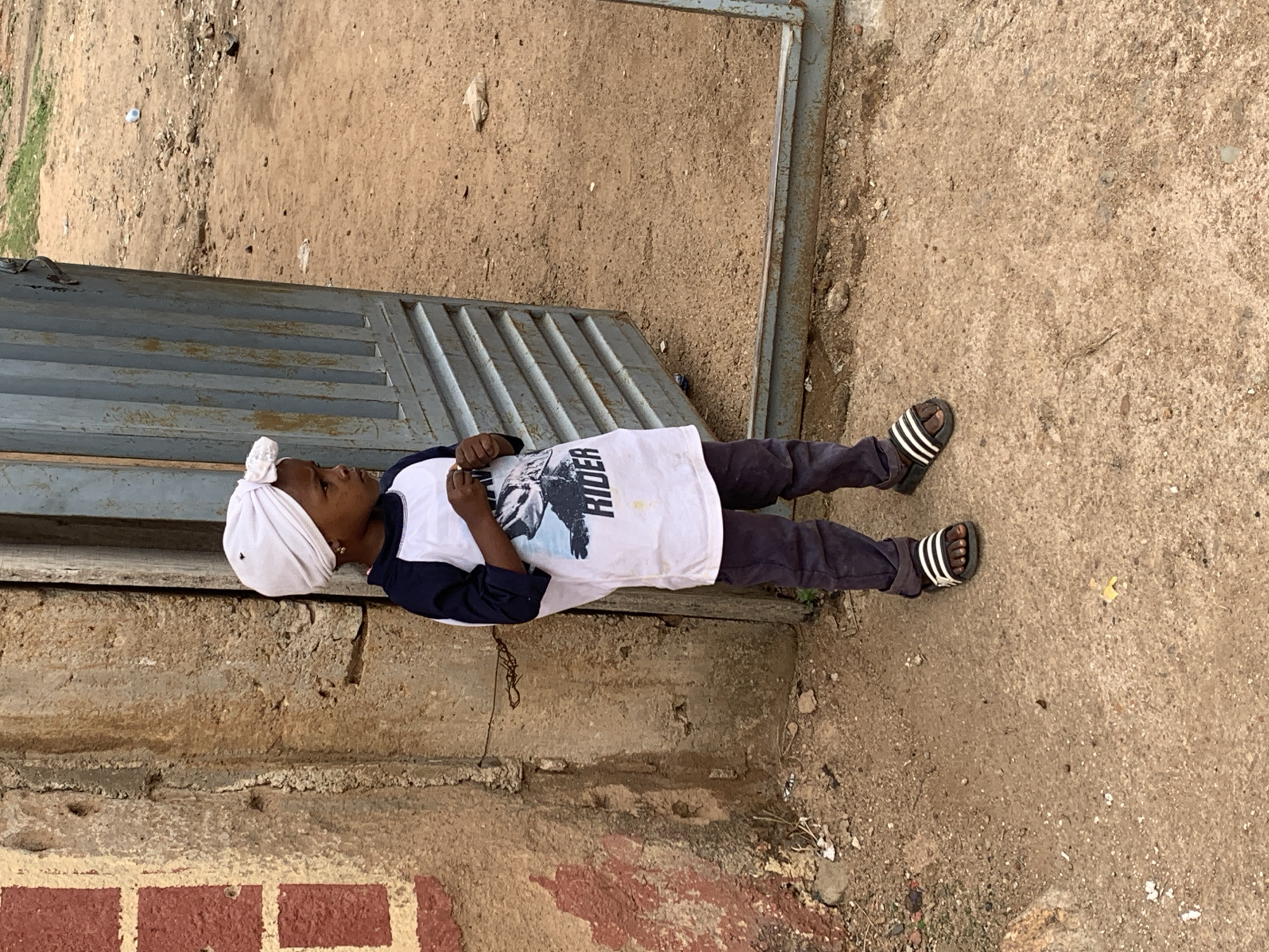 A child lies on a metal grate or low platform next to a wall. The child wears a white head covering, a navy blue and white t-shirt with "Rider" printed on it, dark pants, and black and white striped sandals. The location is in Jos, Nigeria, given the style of the building wall and the surrounding dirt and gravel ground. The grate/platform and wall are positioned beside a dirt/gravel area.