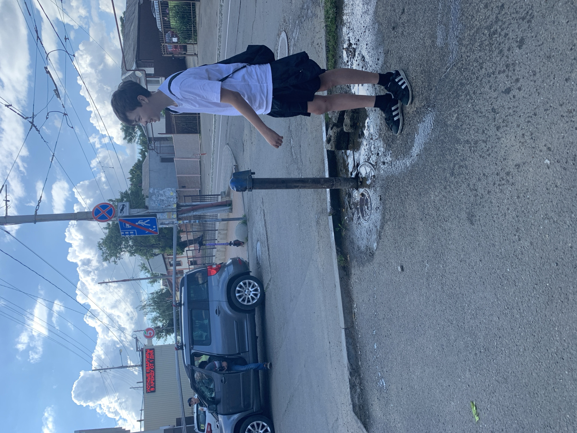 A young male is standing on a wet asphalt surface near a fire hydrant. He is wearing a white t-shirt, black shorts, black socks, and black athletic shoes with white stripes. He has a black bag slung over his right shoulder. Behind him, a dark grey SUV is parked with its driver's side door open. A person is partially visible inside the SUV. The background shows a street scene with buildings, a traffic light, and overhead wires. The sky is cloudy.