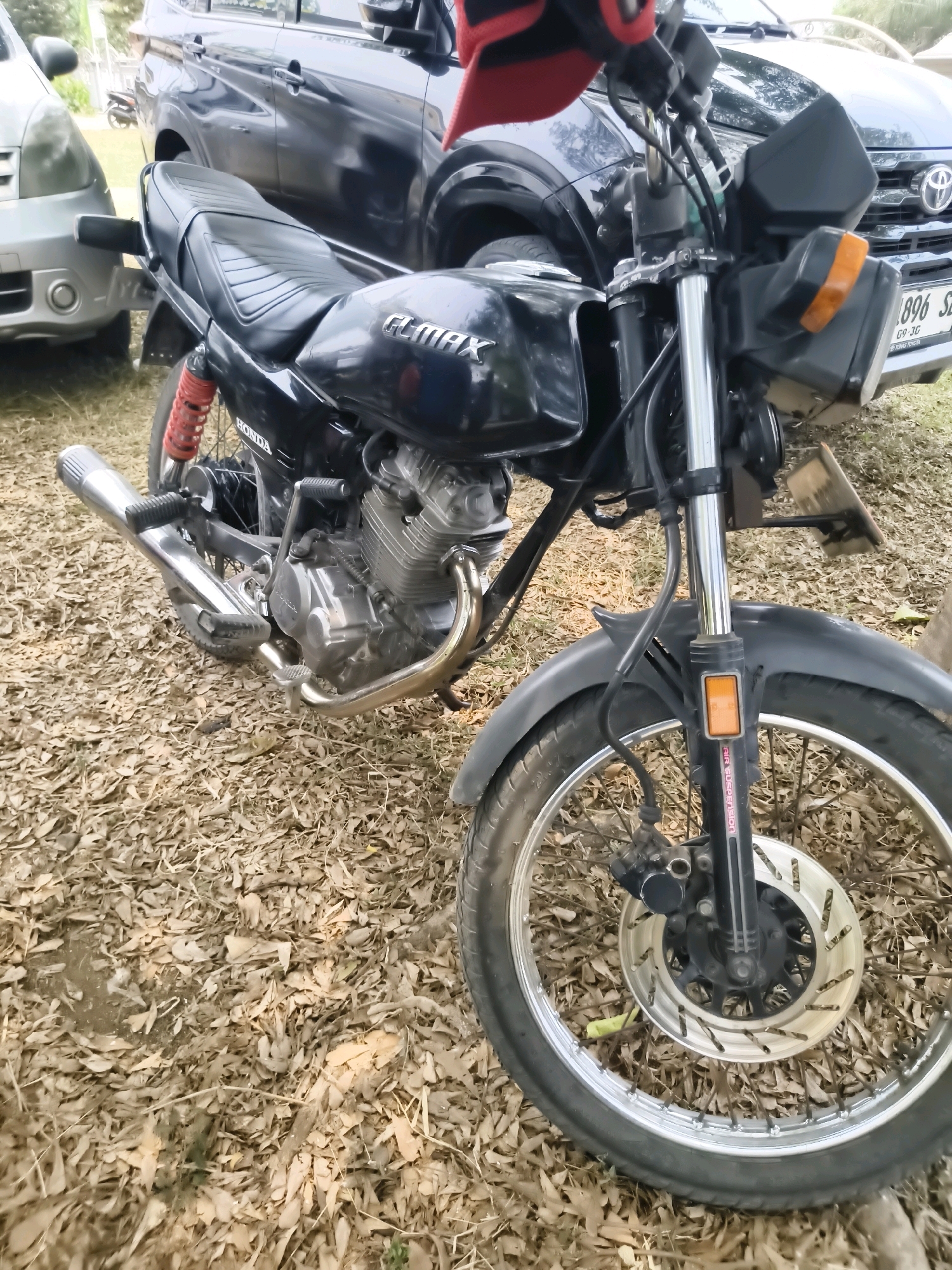 A black Honda motorcycle is parked on a ground covered with dry leaves, in front of a dark colored SUV. The motorcycle has a red rear suspension. A silver car is partially visible on the left side of the image. The license plate of the SUV reads "896 SZ". The scene appears to be outdoors, possibly in a residential area or parking lot in Puloampel, Indonesia.