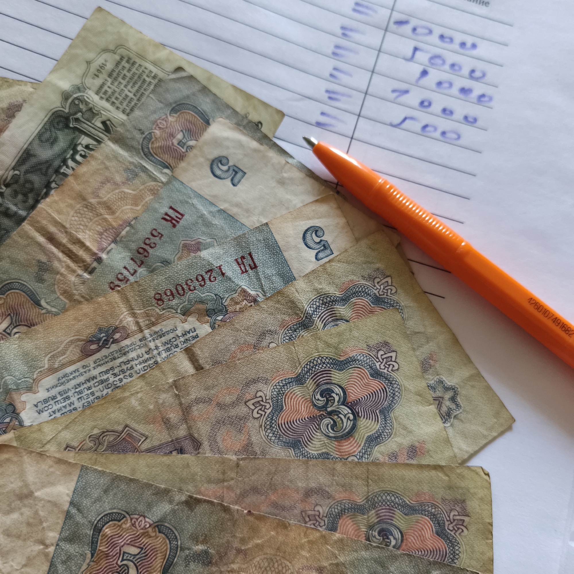 This media file shows a close-up, overhead view of several worn Soviet-era banknotes laid out on a surface that appears to be a lined notebook or ledger. The banknotes are various denominations, with visible numbers "5" and "3" on some. The colors are muted and faded, suggesting their age. An orange pen lies diagonally across the banknotes and the notebook, its tip pointing towards the bills. The notebook has handwritten numbers in blue ink, possibly a list of financial figures, including "500", "1000", and "5000".
Visible text on the banknotes includes "1961", "ГА 1263068", "ГК 5367759", and longer phrases in what appears to be Russian and possibly other languages. Some of this text includes "ПОДДЕЛКА ГОСУДАРСТВЕННЫХ КАЗНАЧЕЙСКИХ БИЛЕТОВ ПРЕСЛЕДУЕТСЯ ПО ЗАКОНУ", which translates to "Counterfeiting state treasury notes is punishable by law." Another visible fragment is "PENKI RUBLIA ЧИНЧ РУБЛЕ- PIECI RUBĻI БЕШ СОМ", which seems to indicate multiple currencies or denominations. A serial number, "4260107491682", is also visible on the orange pen.
The scene is still and focused on the financial items. The lighting is bright, suggesting it might be daytime, indoors, possibly in an office or home setting, given the notebook. There are no people visible in the frame. The overall impression is one of financial record-keeping or perhaps a study of historical currency.