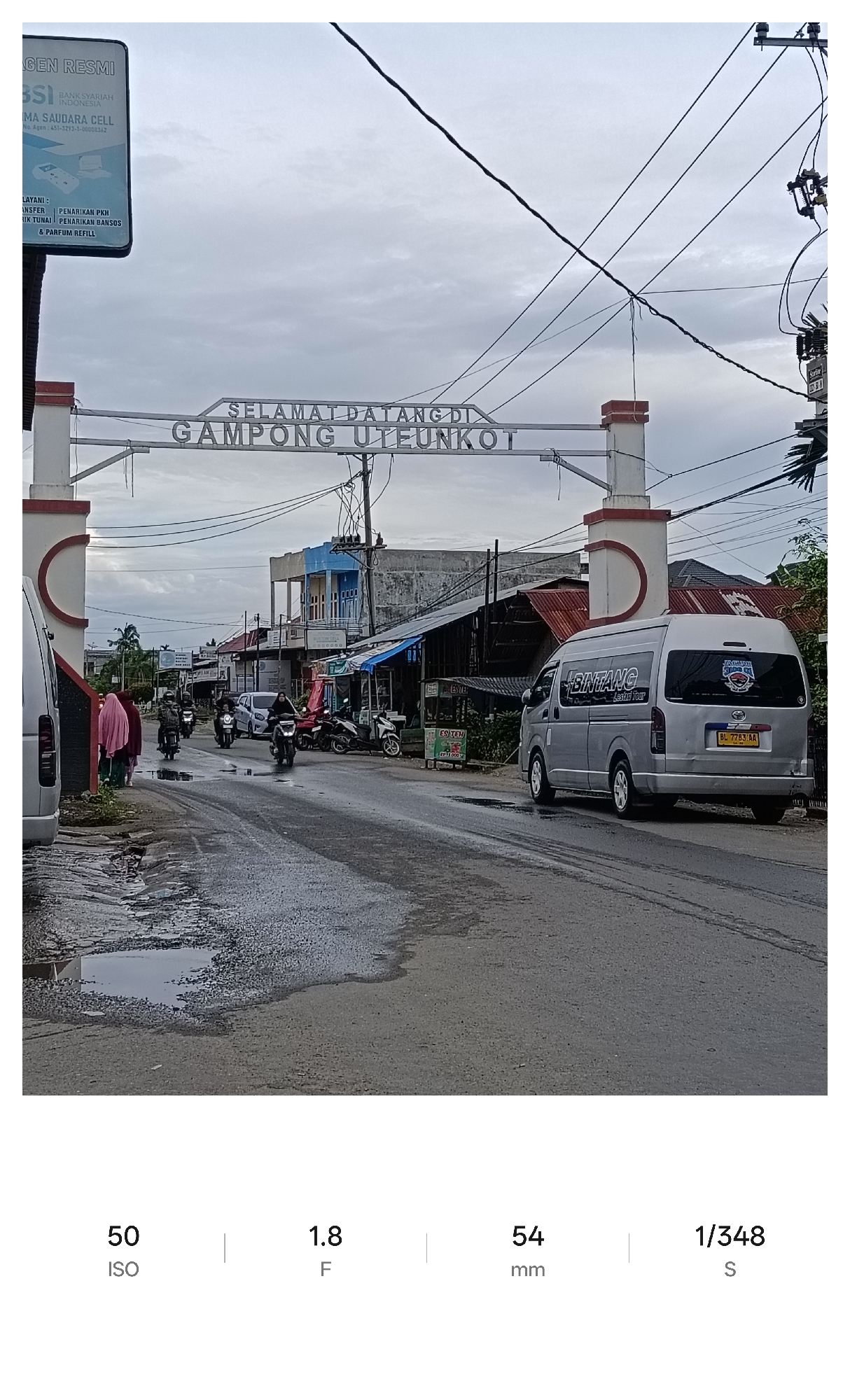 The image depicts a street scene in Gampong Uteunkot, Lhokseumawe, Indonesia. A large archway spans the road, displaying "SELAMAT DATANG DI GAMPONG UTEUNKOT" (Welcome to Gampong Uteunkot) in silver lettering. To the left, a sign reads "GEN RESMI BSI BANK SYARIAH INDONESIA MA SAUDARA CELL" (Official Agent BSI Bank Syariah Indonesia MA SAUDARA CELL). Vehicles, including a silver van with "BINTANG Lestari Tour" written on its side, a few motorcycles, and other cars, are visible on the street. Several people, some wearing pink headscarves, are walking on the left side of the road. Shops and buildings line the street, with one store awning displaying "ES TEH 1000". The road surface is damp with puddles. Overhead, power lines crisscross the sky, which is overcast.