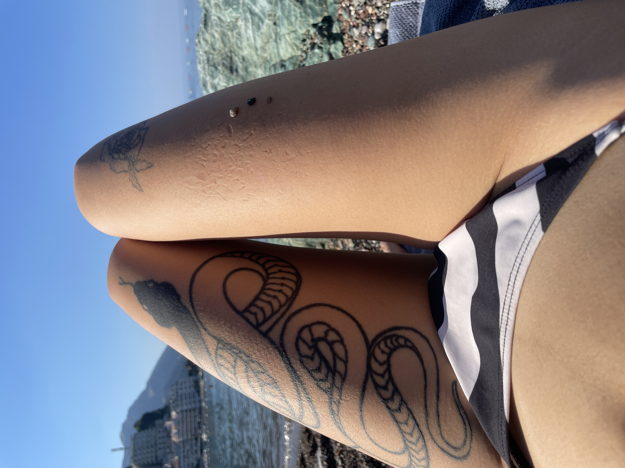 In Rafailovići, Montenegro, a close-up shot focuses on a person's legs, adorned with a snake tattoo on the left thigh and a smaller floral tattoo on the right thigh. The person is wearing a black and white striped bikini bottom. The background reveals a sunlit beach with the sea and a coastal town nestled against a hillside. The water is calm, and the sky is clear blue. Rocks and pebbles are visible on the beach.