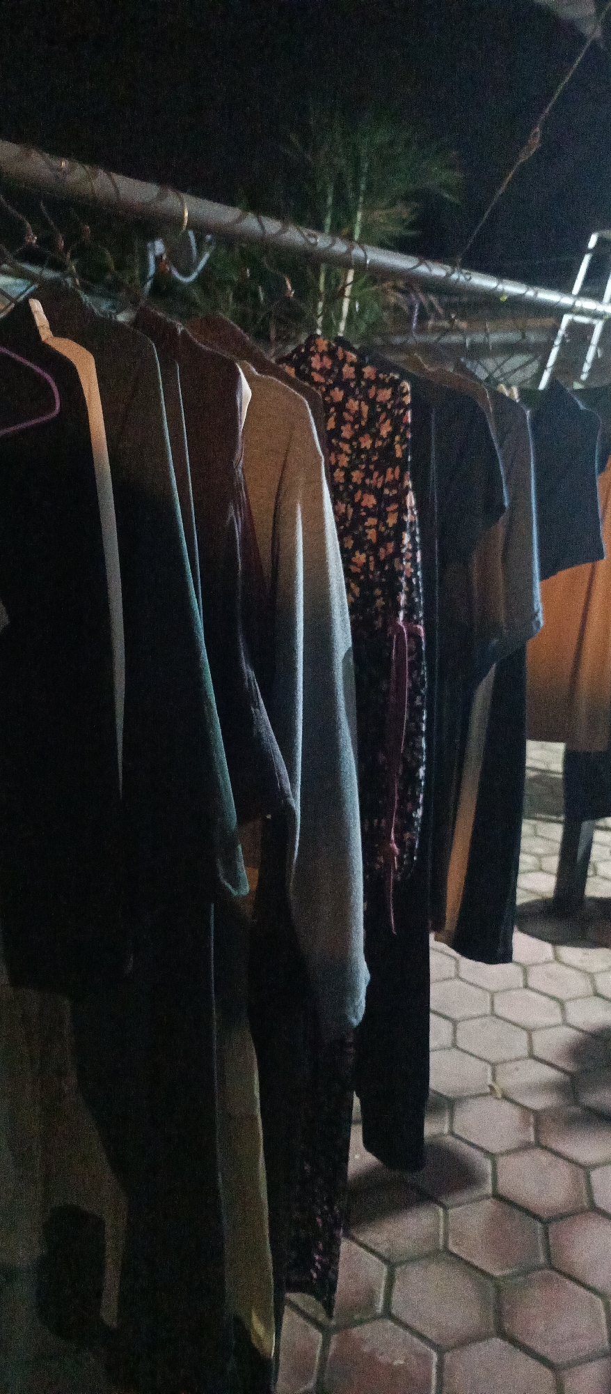 The image depicts a clothesline with several garments hanging outdoors at night. The clothing items include shirts and possibly a dress with a floral pattern. The clothesline is supported by a metal rod. In the background, there are some leafy branches, suggesting the presence of trees or bushes. The ground is paved with hexagonal tiles. The scene appears to be in a residential area, potentially in Arjasa, Indonesia, given the context provided. There are no people visible in the image. The lighting conditions indicate it is dark, with artificial light casting shadows.