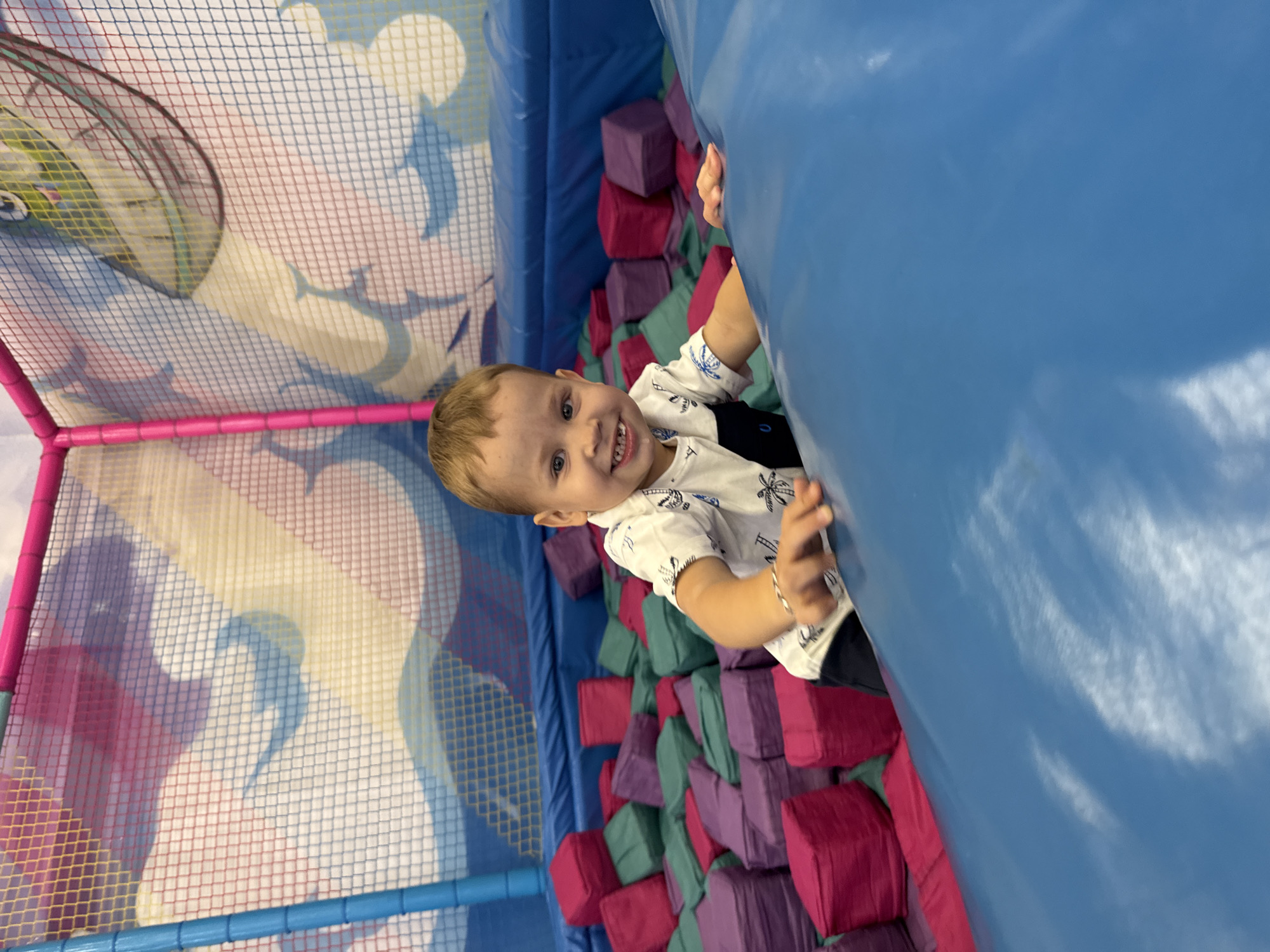 A young boy with light brown hair smiles widely while looking at the camera, his mouth slightly open. He is wearing a white short-sleeved shirt with a dark blue collar and a pattern of palm trees. The boy is positioned in a ball pit filled with brightly colored foam cubes in shades of pink, purple, and green. He is leaning on a large, inflatable blue structure, with his hands resting on its surface. The background consists of a netted enclosure with a colorful mural featuring abstract shapes and what appears to be a cartoon fish. The scene appears to be set within a children's play area. The image was taken in Zhukovo, Russia.