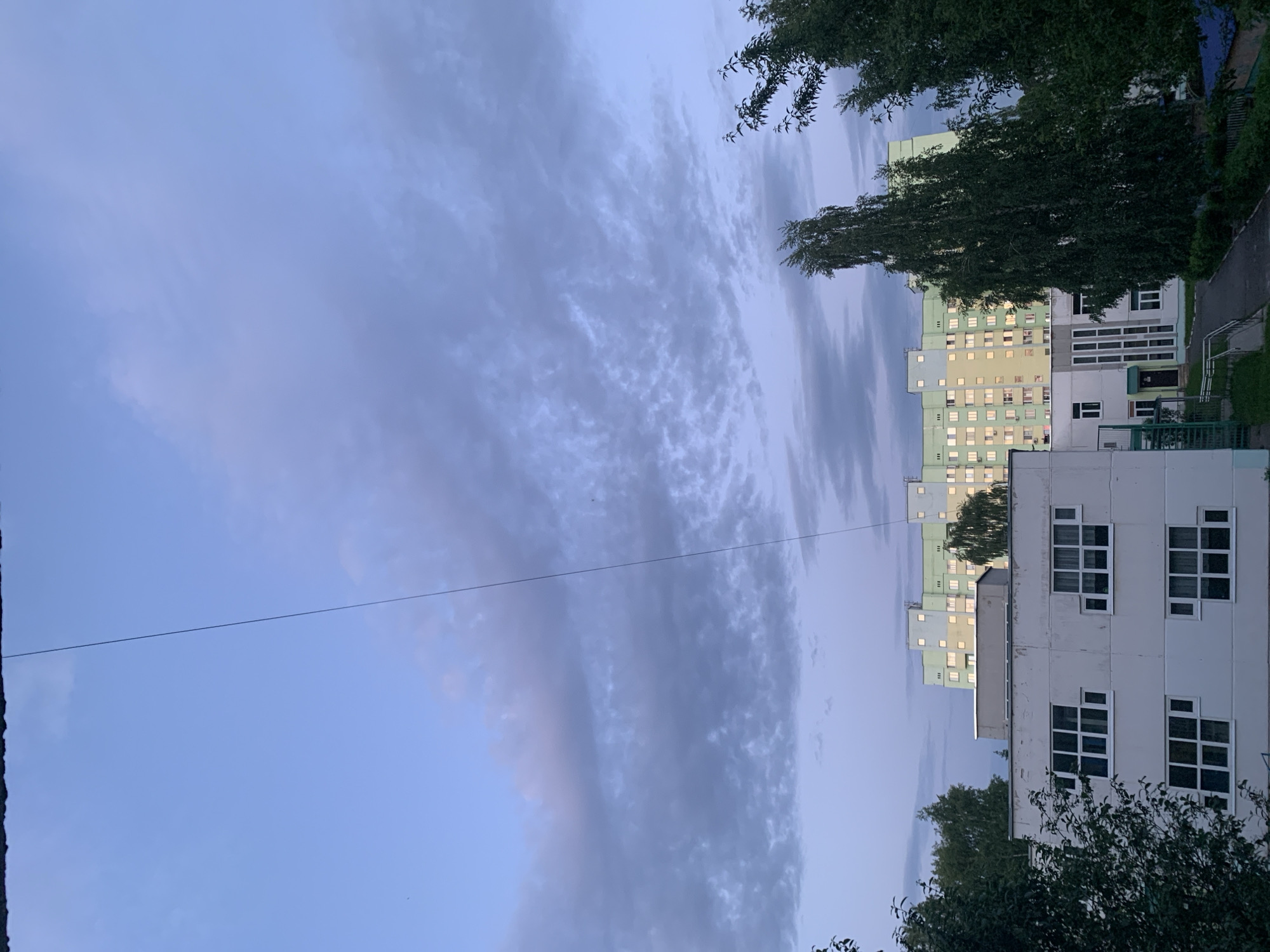 The image depicts a view in the city of Chaikovsky, Russia, at dusk or dawn. It showcases residential buildings with various colors, including white, green, and yellow. Many windows have lights on inside. Trees with green foliage are visible in the foreground and surrounding the buildings. The sky is filled with cloud formations, suggesting a cloudy or overcast day. A thin wire stretches across the upper portion of the frame. The perspective suggests the photo was taken from a low vantage point.