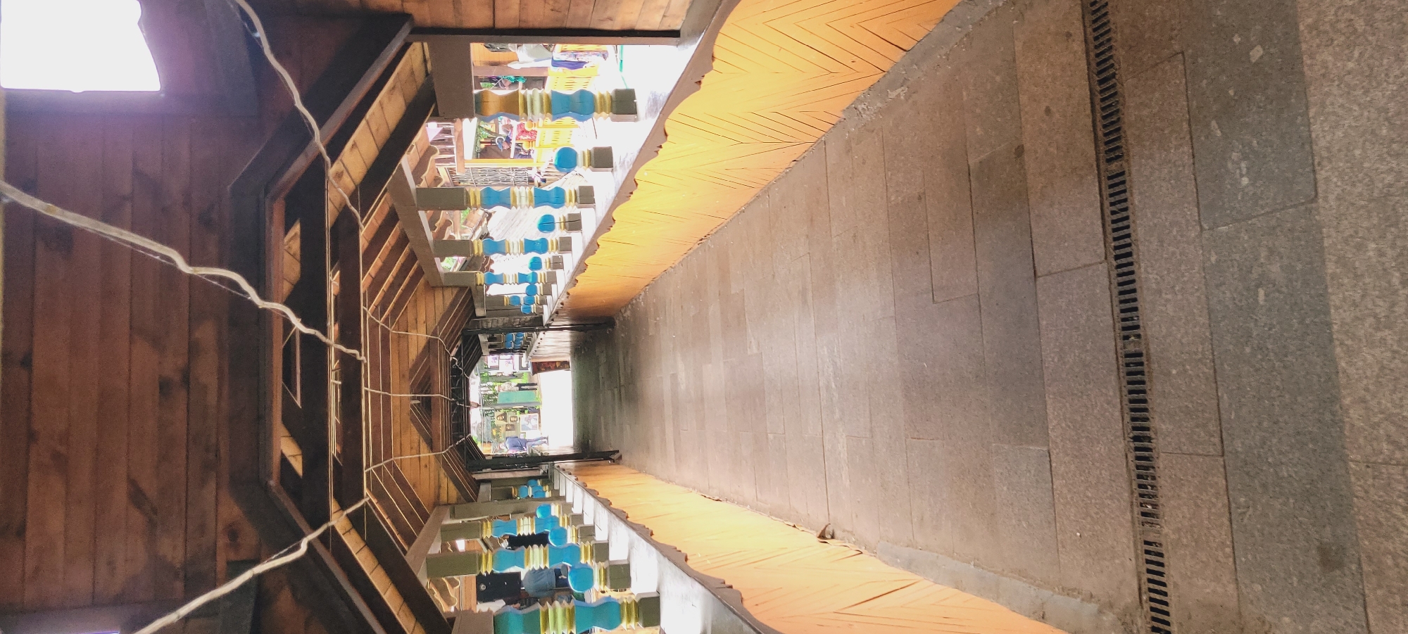 A street-level perspective looking down a narrow pedestrian thoroughfare in Unknown, Unknown. The right side is a paved walkway with a drainage ditch running along its edge. The left side is a building with wooden walls and supports. Overhead, decorations are hung. At the end of the passage, an exit to the street with daylight can be seen.