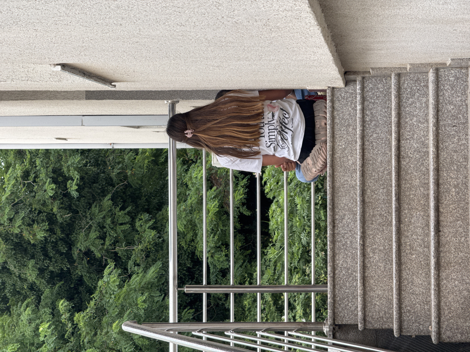The image captures an outdoor scene from a slightly elevated perspective, overlooking dense green foliage. The setting appears to be a balcony or a stairway landing adjacent to a building.
The primary subject is a person, likely female, viewed from behind. They are situated on the left side of the frame, facing away from the camera and towards the lush trees. The person has long, wavy, light brown hair with lighter highlights, which is pulled back at the crown with a small pink scrunchie. They are wearing a white short-sleeved t-shirt with prominent black text on the back that reads "You Are Simply Perfect". Below this, smaller text is visible, stating "Be Brave for Yourself". Part of black lower garments and light-colored, possibly beige or pinkish, sneakers can also be seen.
To the right of the person, a set of light grey, speckled granite stairs descends, featuring prominent vertical lines. A polished silver metal railing, composed of vertical and horizontal bars, extends along the edge of the platform and the stairs. The background is filled with a vibrant, thick expanse of green trees and bushes, suggesting a natural or well-landscaped environment.
Above the person, a textured, light-colored ceiling is partially visible, from which a grey, elongated fixture is mounted. The lighting suggests it is daytime, with soft, ambient illumination. The scene conveys a relaxed, outdoor atmosphere in a location such as Budva, Montenegro.