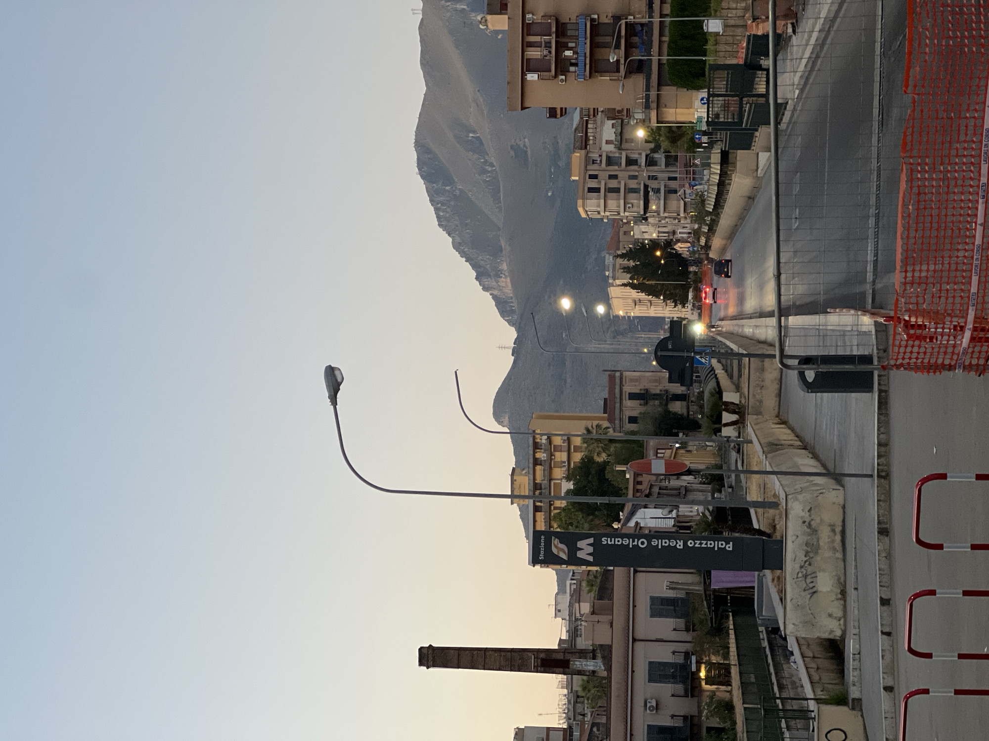 The image depicts a street scene in Palermo, Italy, likely during twilight, with a large mountain range in the background. In the foreground, a road is lined with traffic barriers and construction fencing. A sign indicates "Stazione" and "Palazzo Reale Orleans," suggesting proximity to a train station or transportation hub. Several vehicles are visible on the road, some with their headlights on. Buildings of various architectural styles, including apartment buildings and what appears to be an older industrial structure with a tall chimney, line the street. Streetlights are illuminated, casting a glow on the pavement. The sky is clear and pale blue, transitioning to a warmer hue near the horizon.