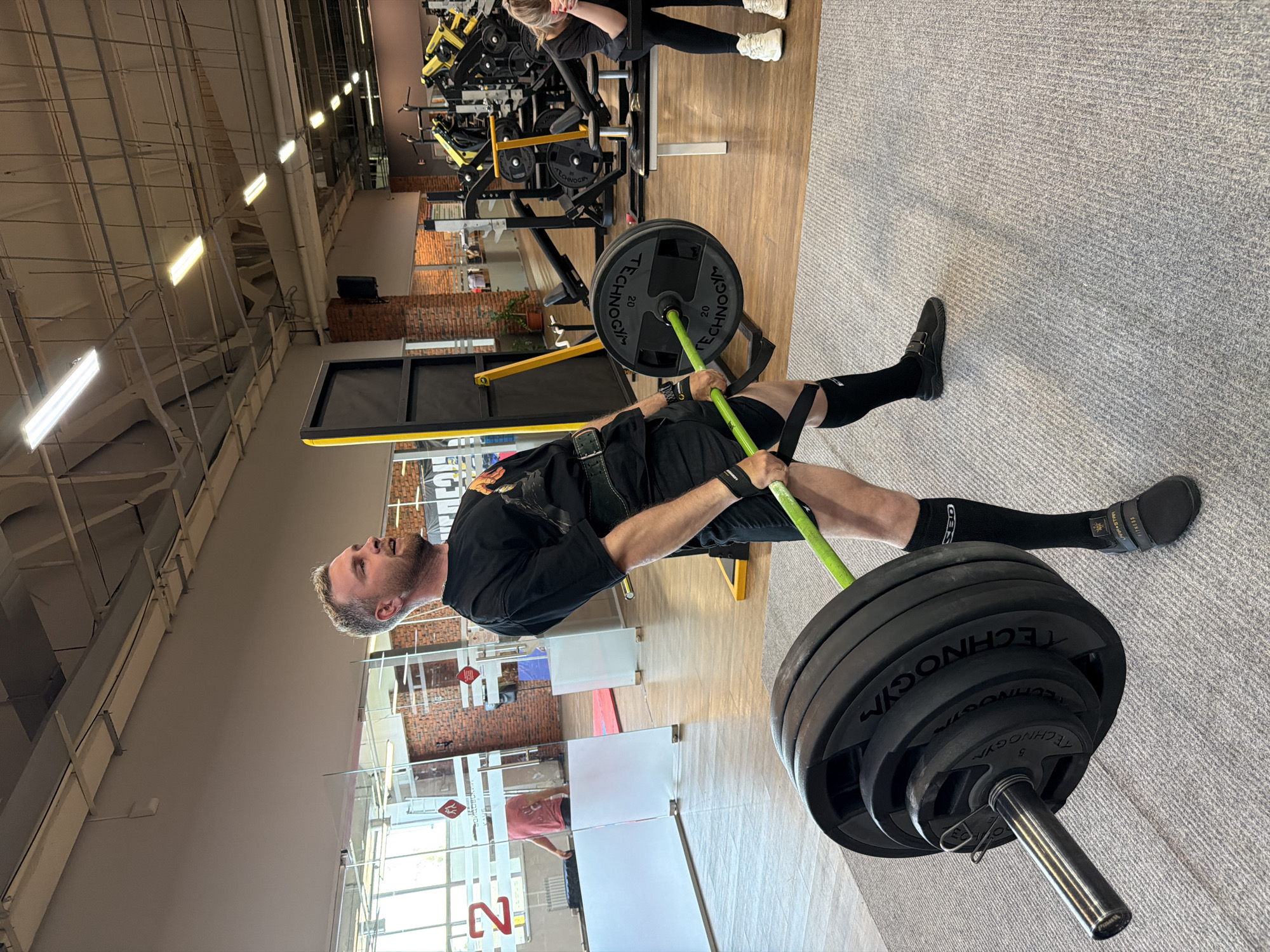 A man with short gray hair and a beard is in a gym, doing a deadlift exercise with a barbell loaded with multiple black weights. The man is wearing a black t-shirt, black shorts, black socks, and black shoes. He also has a weightlifting belt on. The barbell has a green bar and is placed on a rack with a yellow support. The floor is covered in gray carpet. In the background, other gym equipment is visible, along with another person exercising and a brick wall. The gym appears to be well-lit. The image does not provide information to verify the exact location in Chaikovsky, Russia.