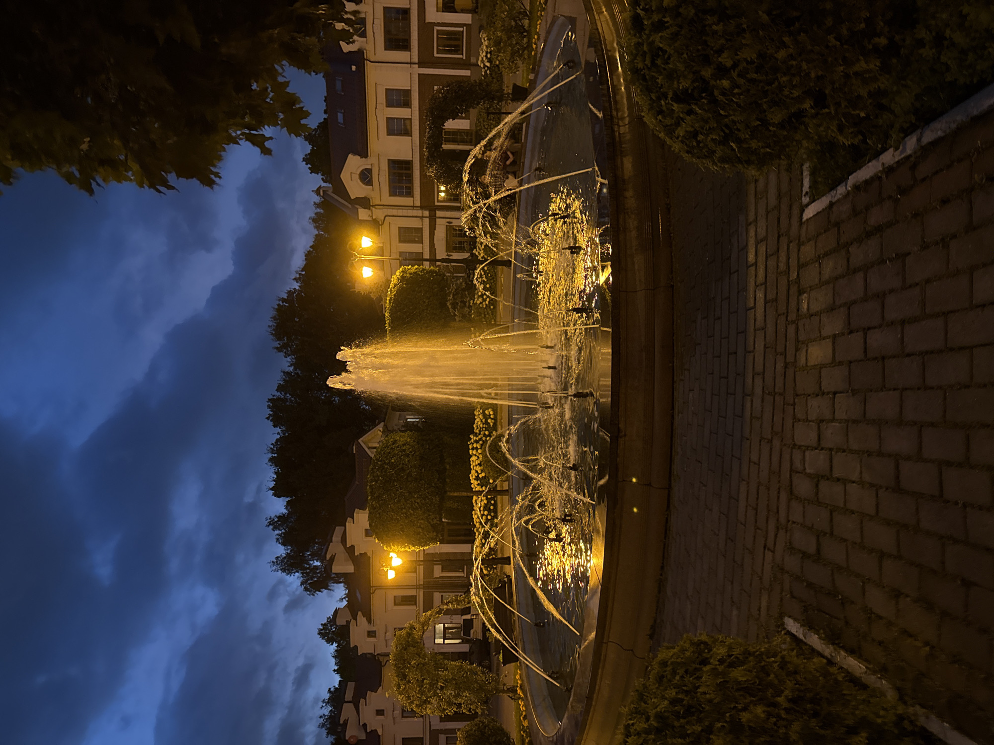 The image depicts an outdoor fountain at night, located in Lopatino, Russia. The fountain is centrally located in a circular basin filled with water, from which multiple jets of water erupt and arc upwards. The water is illuminated by artificial light, casting a golden glow. The surrounding area features a paved walkway made of reddish-brown bricks. On either side of the walkway, there are manicured evergreen bushes. In the background, there are multi-story residential buildings with pitched roofs, characteristic of European architecture. The buildings are painted in light colors, possibly cream or beige, with dark trim around the windows and rooflines. Streetlights with a warm yellow glow are visible between the buildings and behind the fountain. Trees with dense foliage and sculpted bushes are also present in the scene, some creating arched entrances or framing the buildings. The sky above is a deep, dark blue, with scattered clouds, indicating twilight or nighttime. A few figures can be seen sitting on a bench behind the fountain. The overall atmosphere is serene and illuminated by artificial lighting against the dark sky.