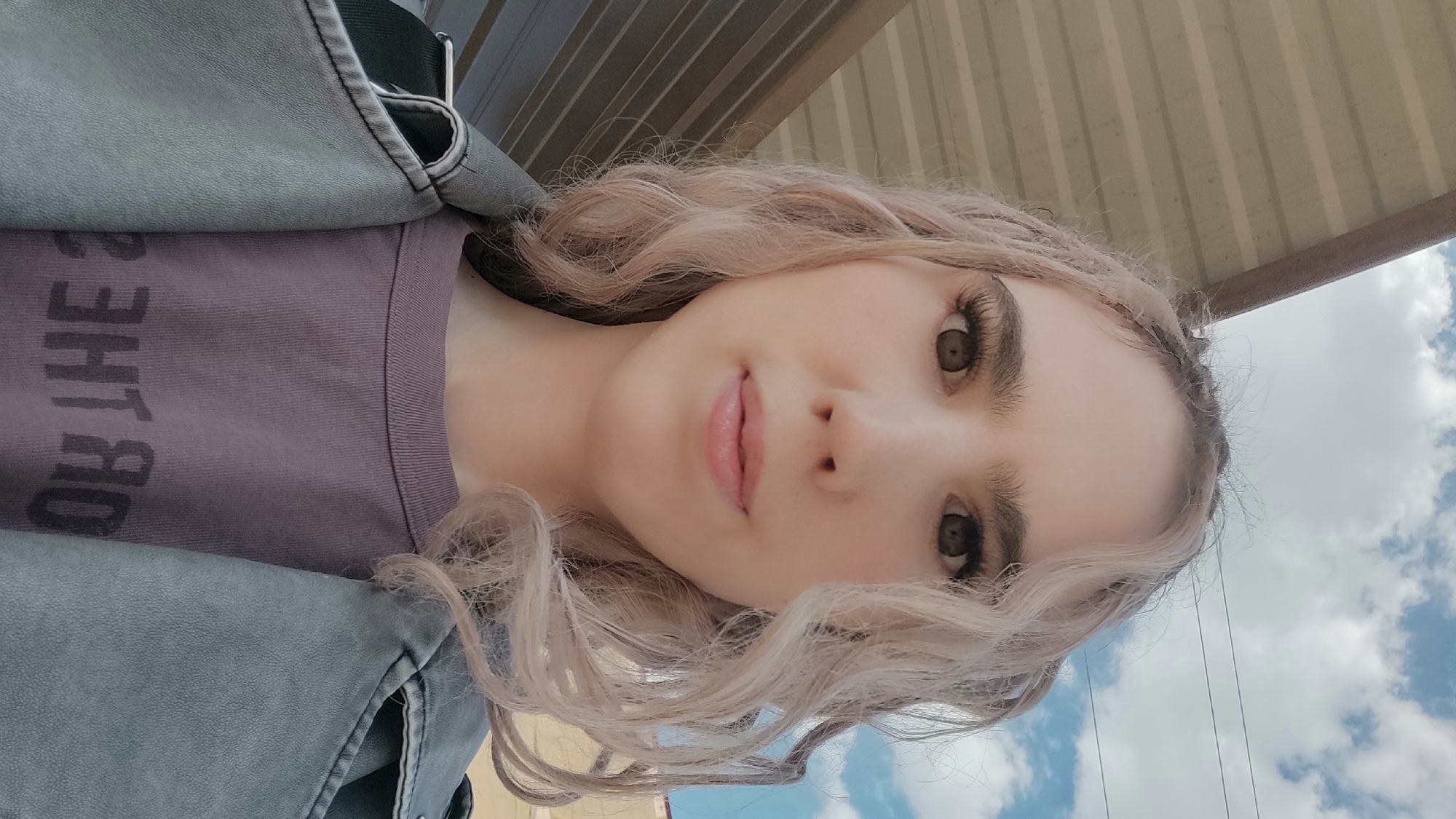 A close-up, slightly rotated image depicts a person with fair skin, light blonde curly hair, and brown eyes. The person is wearing a purple t-shirt with some faded text on it. Over the t-shirt is a gray denim jacket. The background features a corrugated metal awning or roof structure, with a view of a blue sky and clouds visible beyond. There is no information about location in the city of Фоки, Russia, in the image.
