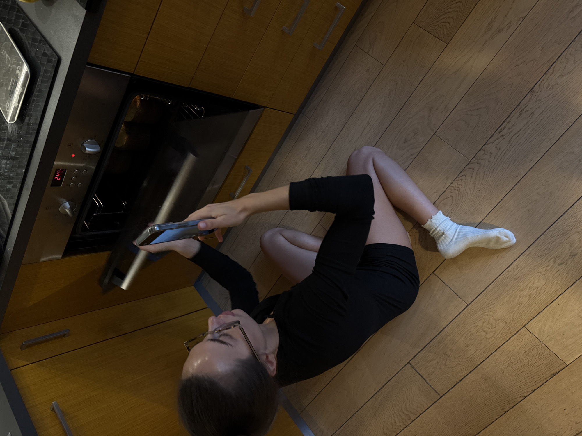 A woman is sitting cross-legged on a wood-paneled floor in a kitchen, facing and holding a mobile phone up to the open door of an oven. She wears a black long-sleeved shirt and black shorts, along with a single white sock on her right foot. Her left foot is bare. She is wearing eyeglasses. The oven is embedded in wood cabinetry with metal pulls. The digital display on the oven shows "2:43". The kitchen appears to be well-lit. There is no indication of the location being Warsaw, Poland, beyond the presence of the oven and associated kitchen features.