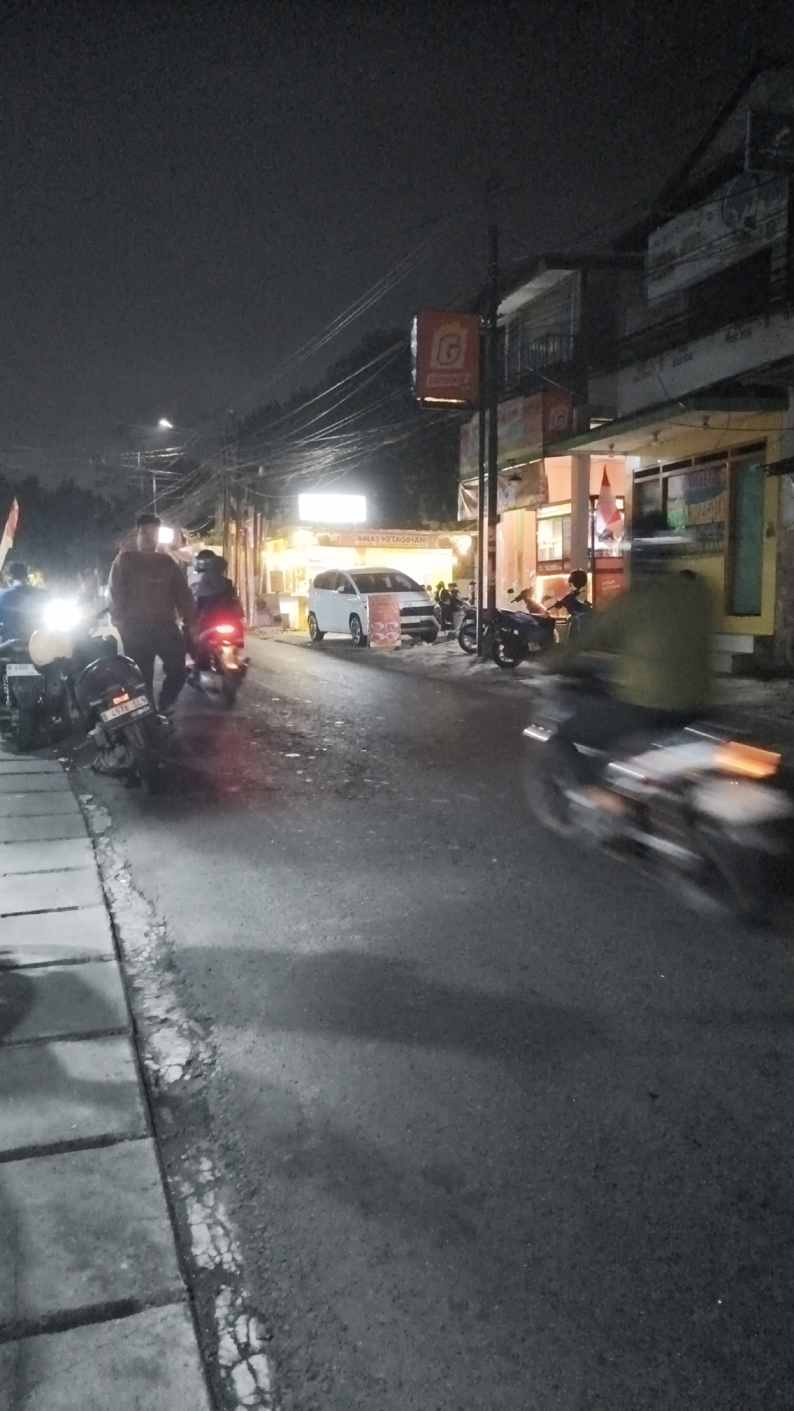 The image depicts a street scene at night in South Jakarta, Indonesia, likely a commercial area with several food stalls or small businesses. In the foreground, a person is riding a motorcycle that is blurred due to motion. The road surface is asphalt with a concrete sidewalk on the left, showing some wear and tear. To the right of the road, there are illuminated stalls and buildings, some with signage. One prominent sign features a red square with a white "G" logo. A white minivan is parked on the side of the road near one of the stalls, which has the text "AWAS KETAGIHAN" visible. Several other motorcycles are parked or being ridden by people. The lighting is from streetlights and the illuminated businesses, creating a nighttime atmosphere with visible shadows. The overall impression is a busy, everyday scene on a street in a populated urban area.