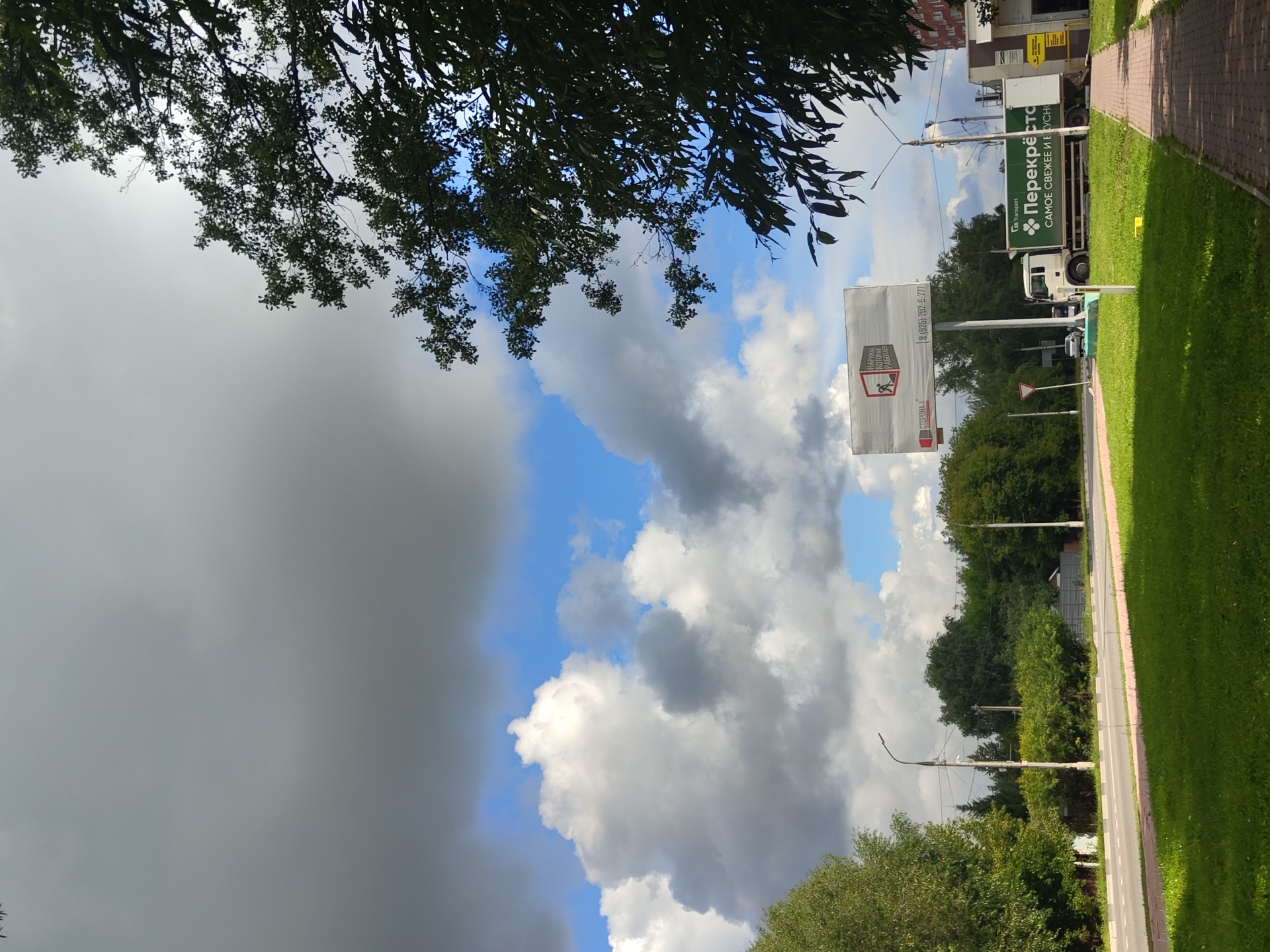 This is a street-level, eye-level shot taken on a cloudy day, showcasing a section of a road in Ramenskoye, Russia. The road, lined with a green grassy median and a paved pedestrian walkway, stretches into the distance. On the right side of the road, a white truck with a green cargo container is parked. The container displays Russian text and a green logo. Adjacent to the truck is a building with a modern facade. To the left of the road, lush green trees and bushes form a natural boundary, with some taller trees on the far left of the frame. In the background, a cluster of buildings, including what appears to be a residential high-rise, peeks through the foliage. A large billboard stands near the roadside, featuring Russian text and a graphic of a worker. The sky is a dramatic mix of grey storm clouds and patches of bright blue, with fluffy white clouds scattered throughout. The overall scene is an everyday urban landscape, with natural elements and man-made structures coexisting.