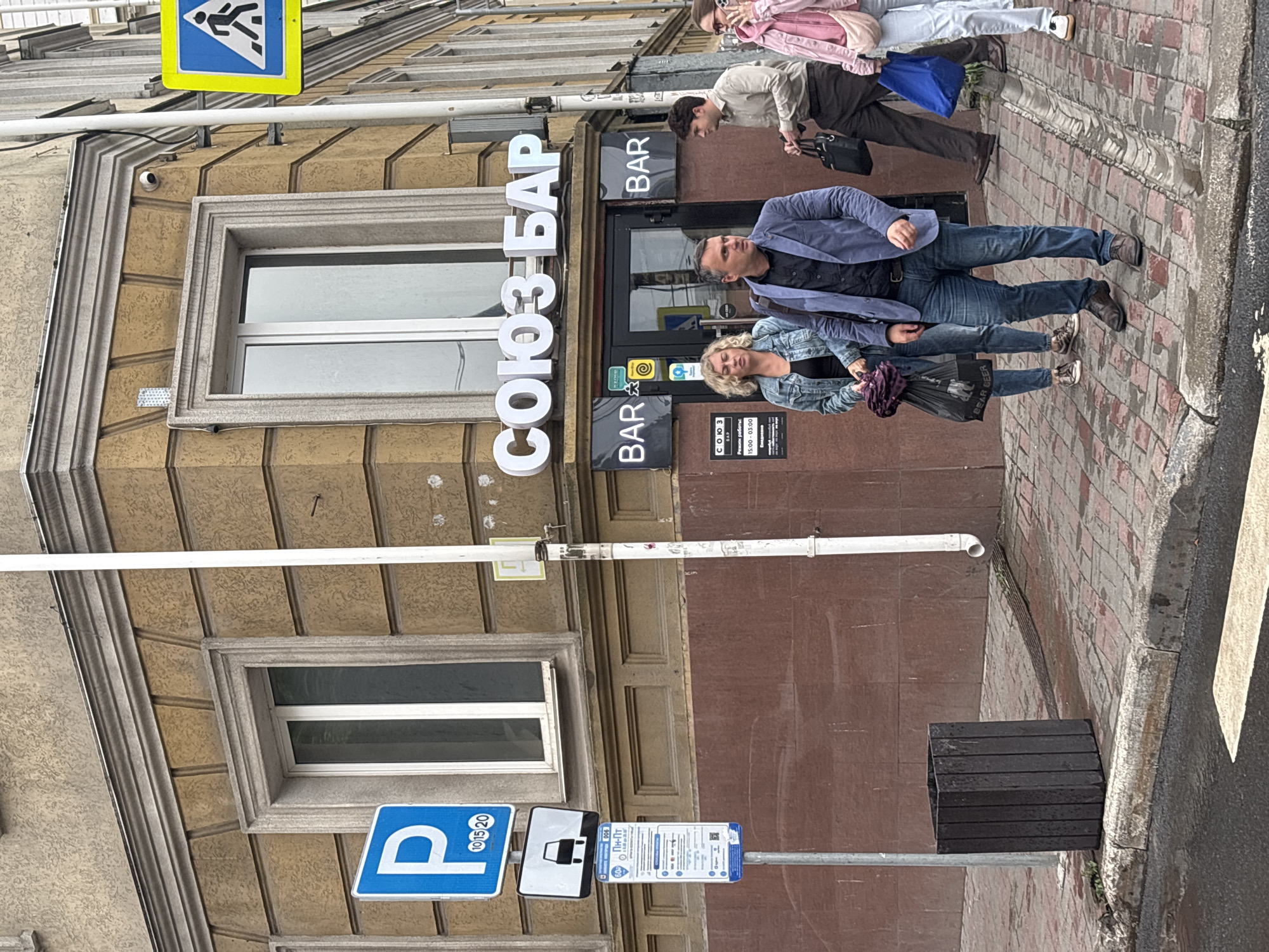 The image shows a street scene in Nizhny Novgorod, Russia. A group of people are standing on a sidewalk in front of a building with a sign that reads "СОЮЗ БАР". The building appears to be a bar or restaurant. There are several signs on the sidewalk, including a parking sign and a bus stop sign. The ground is wet, suggesting it may have recently rained. The people in the image are dressed in casual clothing and appear to be waiting or walking. One person is holding a shopping bag, and another is talking on their phone. The overall atmosphere is that of a typical urban street.