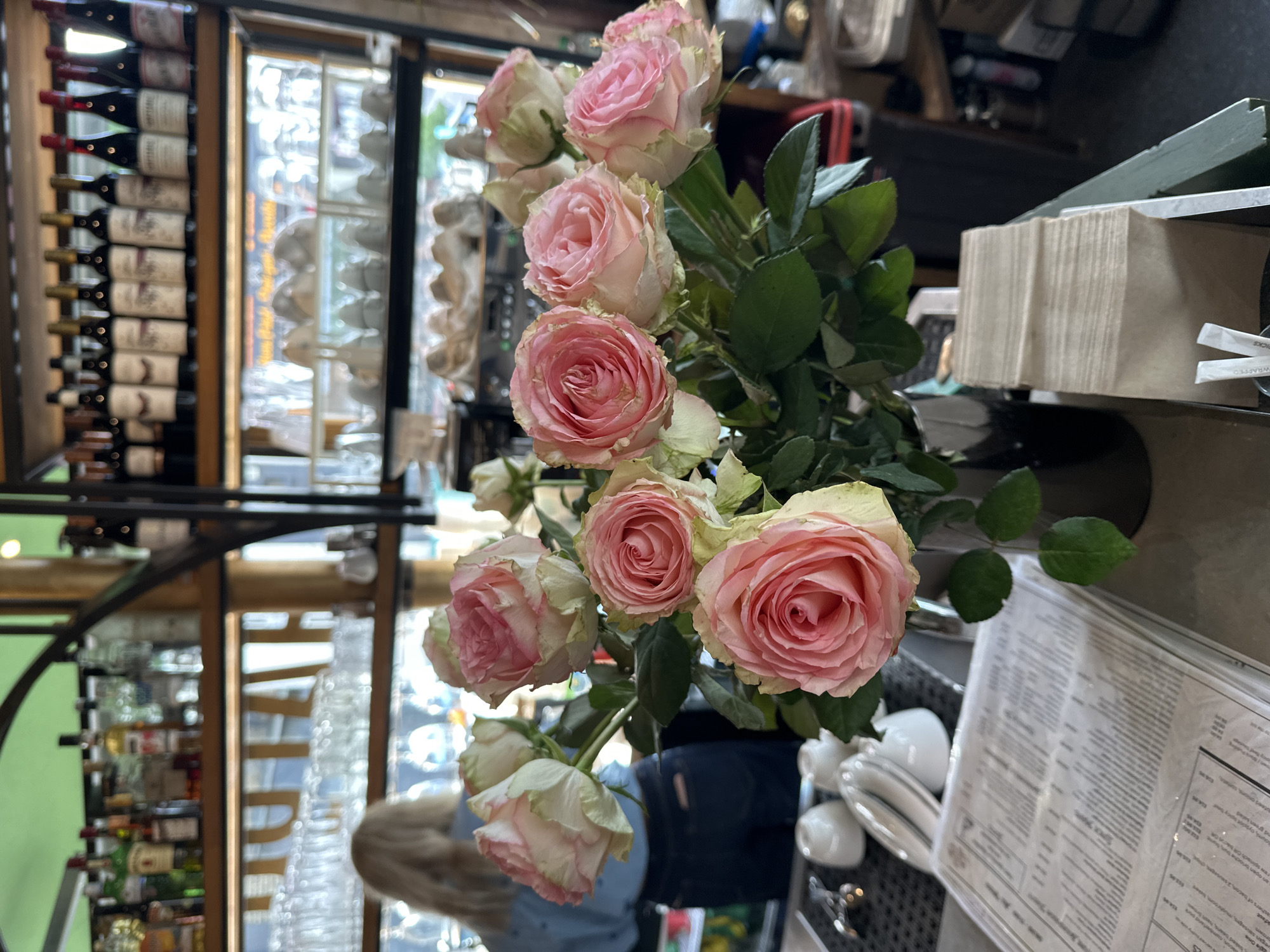 The image presents an indoor scene, possibly a restaurant or bar, with a vase of pink roses as a focal point. The roses are in full bloom, displaying a soft pink hue. Behind the roses, a well-stocked bar is visible, featuring rows of wine bottles and decorative glassware. The environment suggests a sophisticated setting. There is a person partially visible behind the roses. A menu and various items are on the table next to the roses. Without further details, it is not possible to confirm the location in the city of London, United Kingdom.