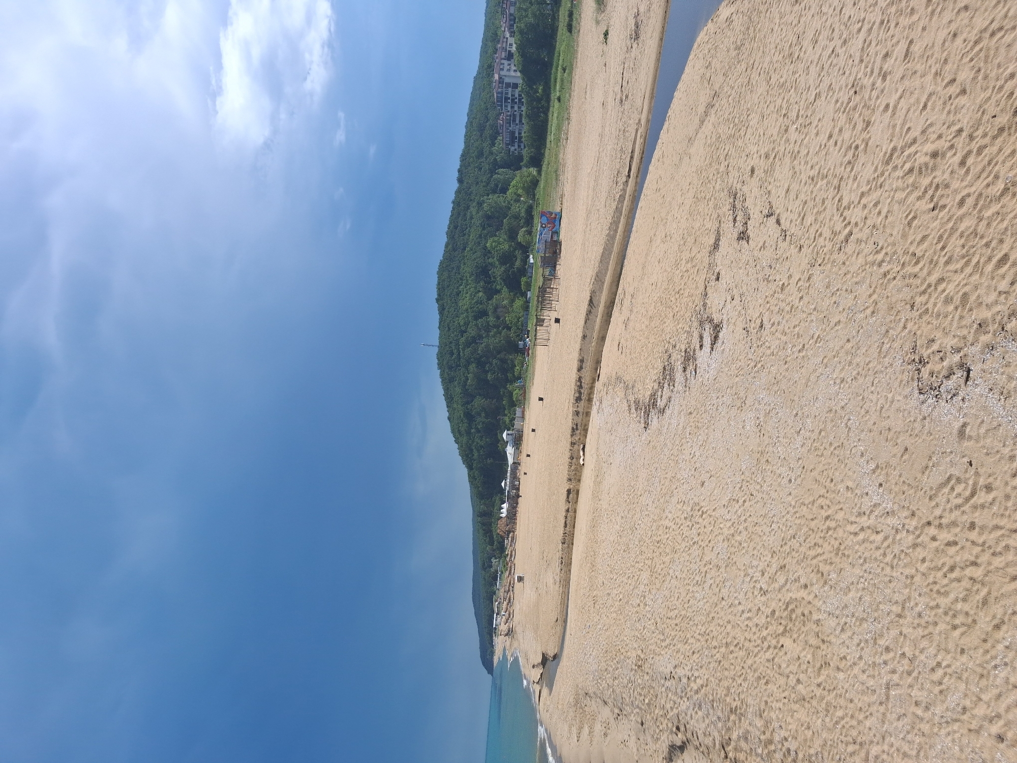 This wide-angle image depicts a coastal landscape in Shkorpilovtsi, Bulgaria, under a partially cloudy sky that shifts from a darker blue on the left to a lighter blue with diffuse white clouds on the right. The turquoise sea occupies the far left of the frame, where gentle waves meet an expansive, light beige sandy beach. A narrow, dark blue stream or river flows across the sandy terrain, paralleling the coastline before curving to discharge into the sea. The beach sand shows distinct ripple patterns and contains scattered organic debris and shell fragments. In the background, a prominent hill densely covered with dark green trees extends horizontally. Along the base of this forested hill, on the beach, several scattered structures with white roofs, potentially beach facilities, are visible. Further to the right, a series of bare wooden frames stands on the sand, adjacent to a brightly colored, multi-paneled sign. On the forested hillside toward the right, several light-colored, multi-story buildings, indicative of residential or hotel complexes, are partially visible amongst the trees. A tall, slender white tower, likely a telecommunications mast, is situated atop the hill, to the left of the buildings. No human figures are present in the scene.