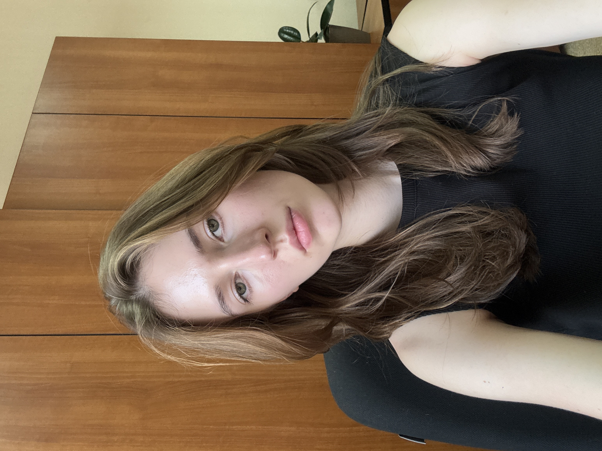 A young woman with long, wavy brown hair and light skin is sitting in an office. She is wearing a black sleeveless top. Her head is tilted slightly to her left. The background consists of a wooden cabinet with two doors and a beige wall. A potted plant is visible on a surface to the right of the woman. The image is a selfie taken from a high angle. The location of this office is not specified in the image or its metadata.