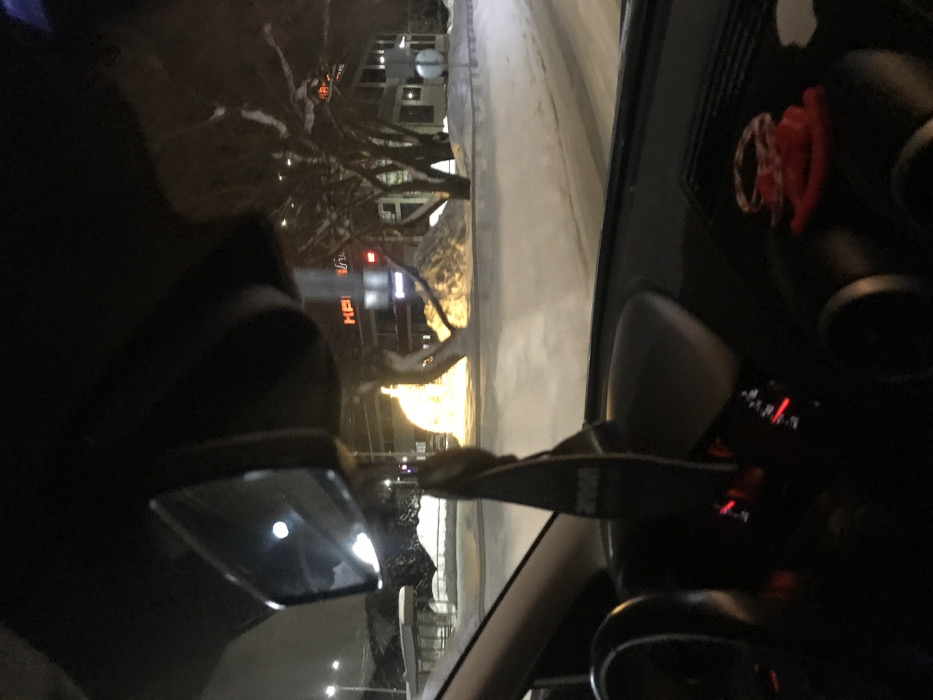 The image presents a nocturnal winter scene observed from the interior of a vehicle in Kremyonki, Russia.
Inside the vehicle, a dark, rectangular air freshener, potentially branded "AVON," hangs from the stem of a rearview mirror, which reflects a circular light source. A segment of a dark steering wheel is visible on the lower left. The dashboard displays red-illuminated gauges and, on its right side, a small red coiled object.
The exterior landscape is entirely covered in snow. The road surface appears partially cleared, contrasting with deep snow accumulation on the verges and sidewalks, forming several mounds. In the mid-ground, a large, spherical or dome-shaped decorative light installation emits a bright, warm yellow glow. Behind this, bare deciduous trees are heavily laden with snow. A multi-story building features red illuminated Cyrillic signage, prominently displaying "КЕ," likely part of "КЕНТАВР" (KENTAVR), a common name for commercial establishments. Another building with illuminated windows is visible further right. A snow-covered bus stop shelter is present on the far left, and a circular road sign is partially visible on the right side of the road. The scene is illuminated by ambient streetlights. No individuals are discernible.