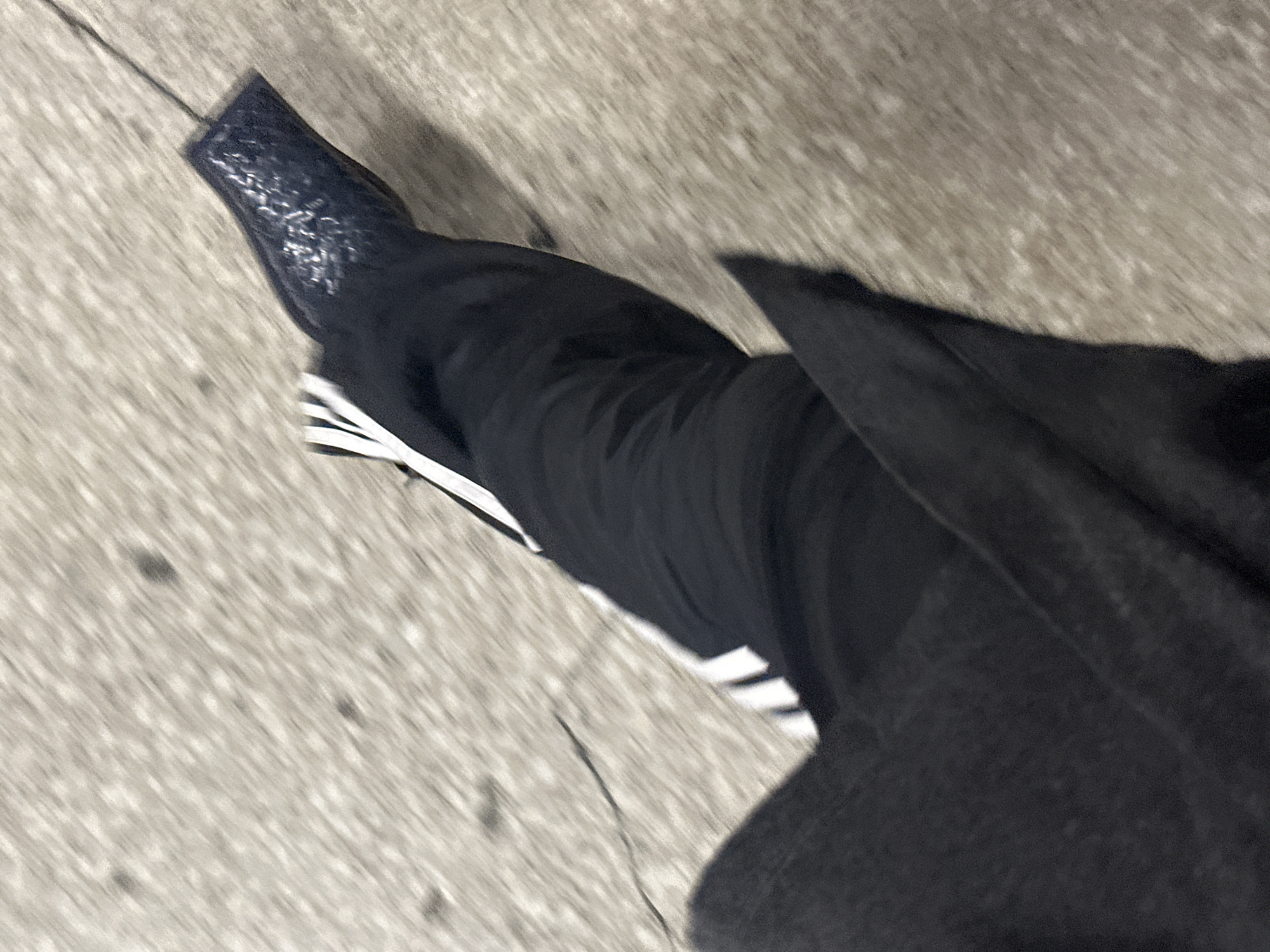 A person's leg is seen from above, with an Adidas shoe with black and white stripes visible. The person is also wearing black pants and a dark overcoat. They are standing on a concrete surface. There is no clear indication of the image being taken in London.