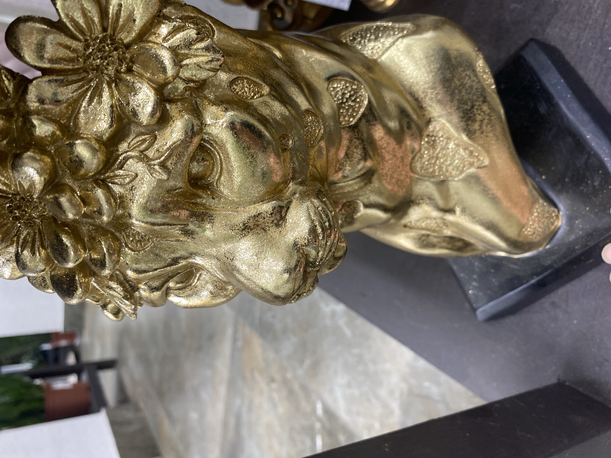 A close-up, angled shot features a golden statue resembling a lion with floral decorations. The statue is mounted on a dark, possibly marble, base. The statue's head is adorned with several flowers in full bloom, and textured patterns mark the lion's body. The statue is indoors with a blurred background suggesting a room or commercial space. Elements of flooring, furniture, and possibly greenery can be seen in the background. There is no evidence to suggest that the image was taken in Yekaterinburg, Russia.