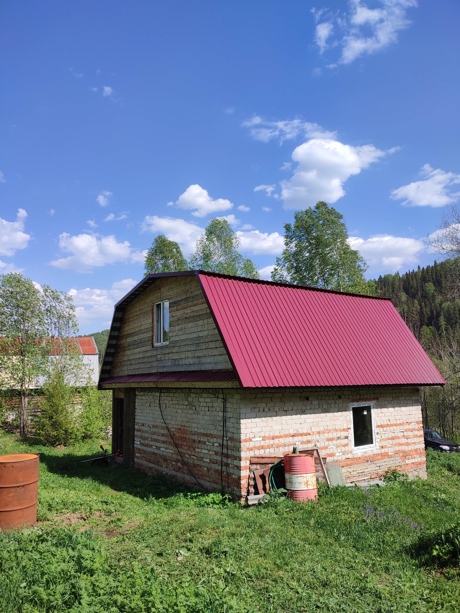 The image shows a two-story building with a red metal roof in Vil'va, Russia. The lower story is made of brick, and the upper story is made of wood siding. A single window is visible on each story. The building appears unfinished or in disrepair, with debris and various objects surrounding it. Two rusty metal barrels sit in the overgrown grassy yard. Trees and a blue sky with scattered clouds form the background. A section of woodland borders the property. The environment suggests a rural or semi-rural location.