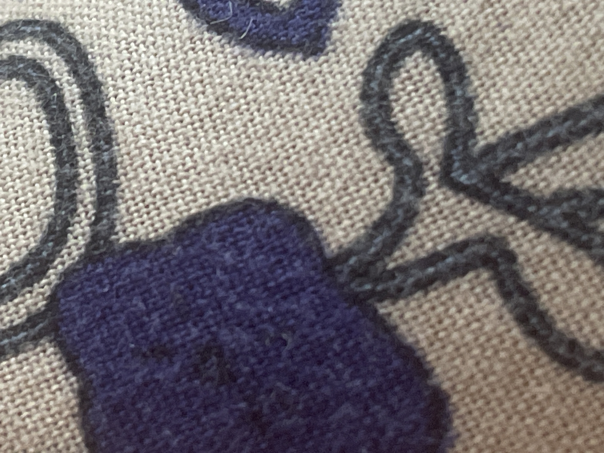 The image displays a close-up, abstract view of a textile with a woven pattern. A large, irregular shape of dark blue fabric is prominent on the left side. This blue fabric appears to be stitched or appliqued onto a lighter, textured background fabric that has a neutral, beige or light brown hue with a visible weave. Overlaying both fabrics are dark, thick, thread-like lines that form curved and looping patterns. These lines are uneven and appear to be embroidered or printed onto the fabric. The lighting suggests a soft, diffused light source, and the focus is on the texture and pattern of the material itself. There is no discernible geographical context, and the image does not depict any elements that would allow for identification of a location in Tashkent, Uzbekistan, or any other city. The scene is entirely focused on the details of the textile.